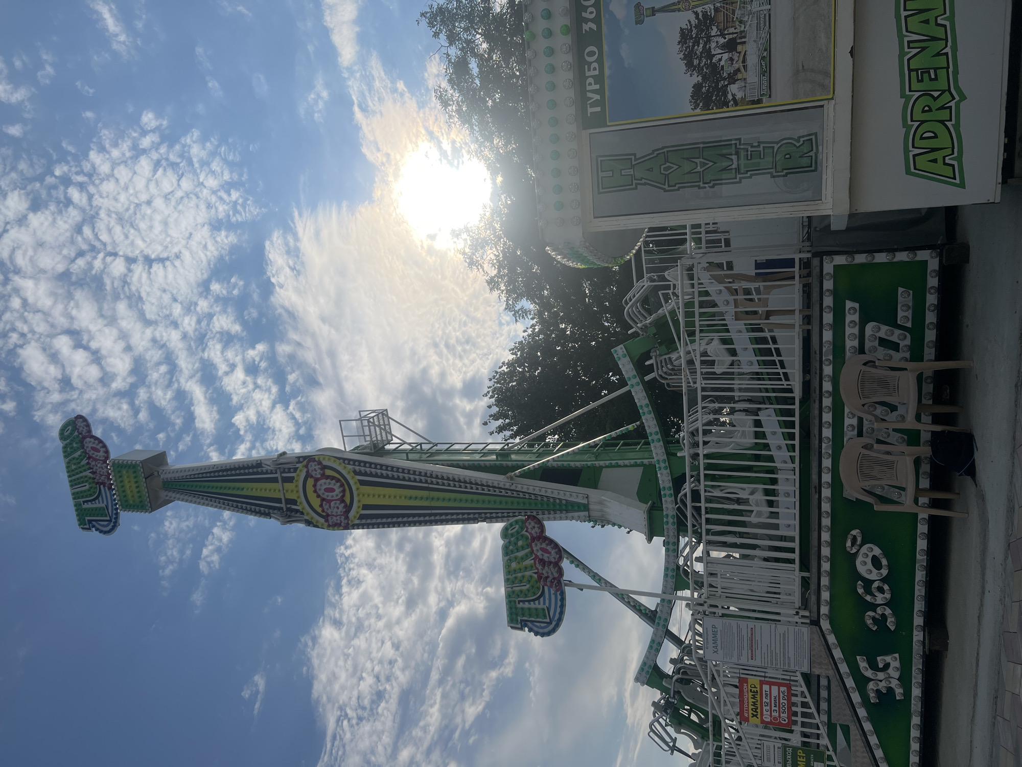 An amusement park ride, identified as "Turbo 360 Hammer," is the central subject of this outdoor photograph, taken during daylight hours in Golubitskaya, Russia. The ride is a towering, multi-colored structure with green, white, and yellow accents, featuring a large vertical arm that swings riders. The sky above is blue with scattered white clouds, and the sun is partially obscured by clouds, casting a bright glare. The base of the ride is a platform with white railings and signage. One sign displays "ATTRAKTION HAMMER" in Russian, along with age and price restrictions. Below the ride's structure, the words "3G 360°" are emblazoned in large green letters. Two empty beige plastic chairs and a dark backpack are placed on the ground in front of the ride. The scene appears to be set within an amusement park or fairground, with trees visible in the background.