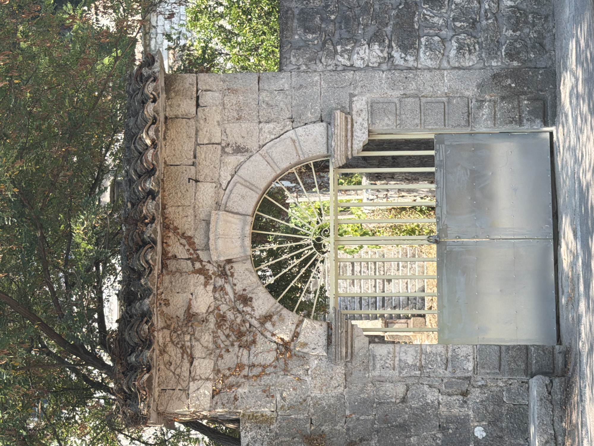 The image shows a weathered stone gateway with a large, plain metal gate in the foreground. Above the gate, an arched metal grate with a radial pattern is visible, framed by carved stone. The stone wall surrounding the gateway is covered in climbing vines with dry leaves. The roof of the structure is made of terracotta tiles, partially obscured by lush green tree branches. Beyond the gate, stone steps ascend into a shaded area, suggesting an entrance to a property or garden. The architectural style appears to be old and robust, typical of coastal Mediterranean regions. The scene is likely located in Prčanj, Montenegro, a town known for its stone architecture and historical buildings along the Bay of Kotor. The lighting suggests it is daytime, with sunlight casting shadows on the ground in front of the gate. No people are present in the image.