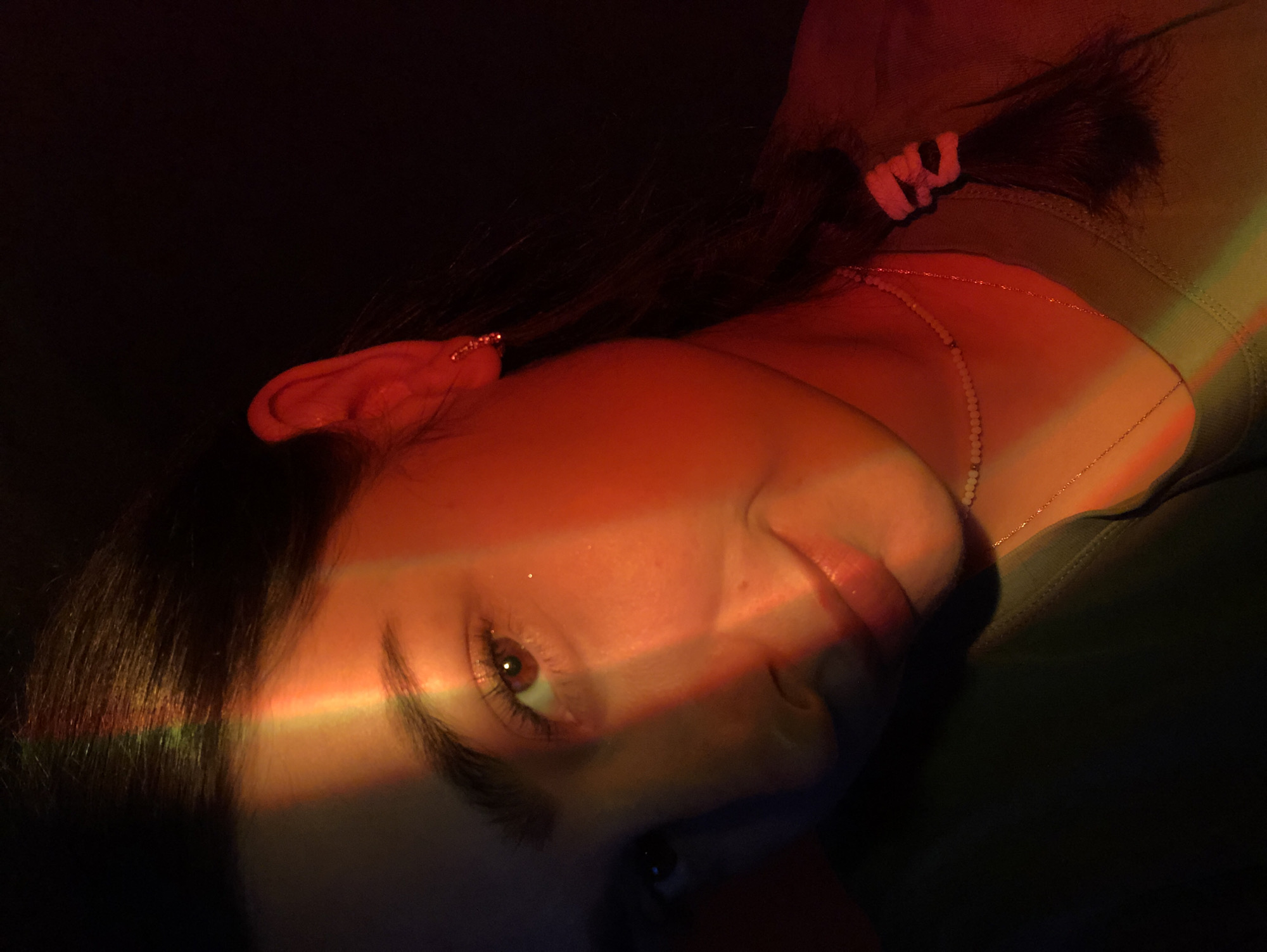A medium close-up photograph shows a young woman against a dark background. She is looking towards the left side of the frame with a slight smile. The lighting creates a rainbow-like effect across her face, highlighting her right eye and cheekbone, and casting red light on her ear and the side of her head. She has dark hair pulled back into a braid. She is wearing a necklace and a green shirt. The image does not provide any information about the city of Noviy, Russia.