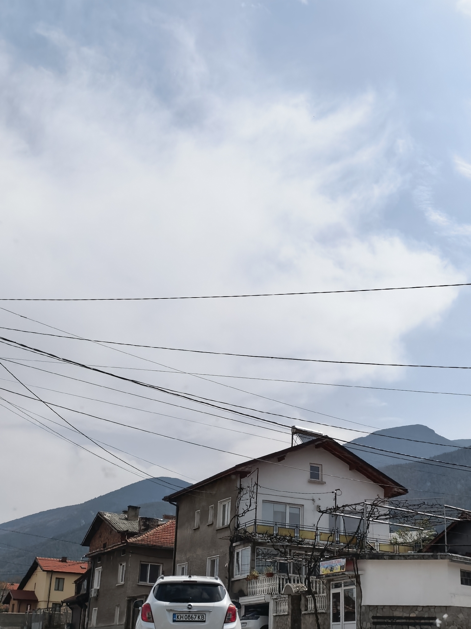 A street scene in the city of Unknown, Unknown, shows a white Opel automobile parked in front of two-story buildings. The automobile, bearing the license plate KH 0867 KB, is positioned on the left side of the image. Buildings with tiled roofs and visible architectural features are present. In the background, a mountain range can be seen under a partly cloudy sky. Overhead, a series of power lines stretch across the view.