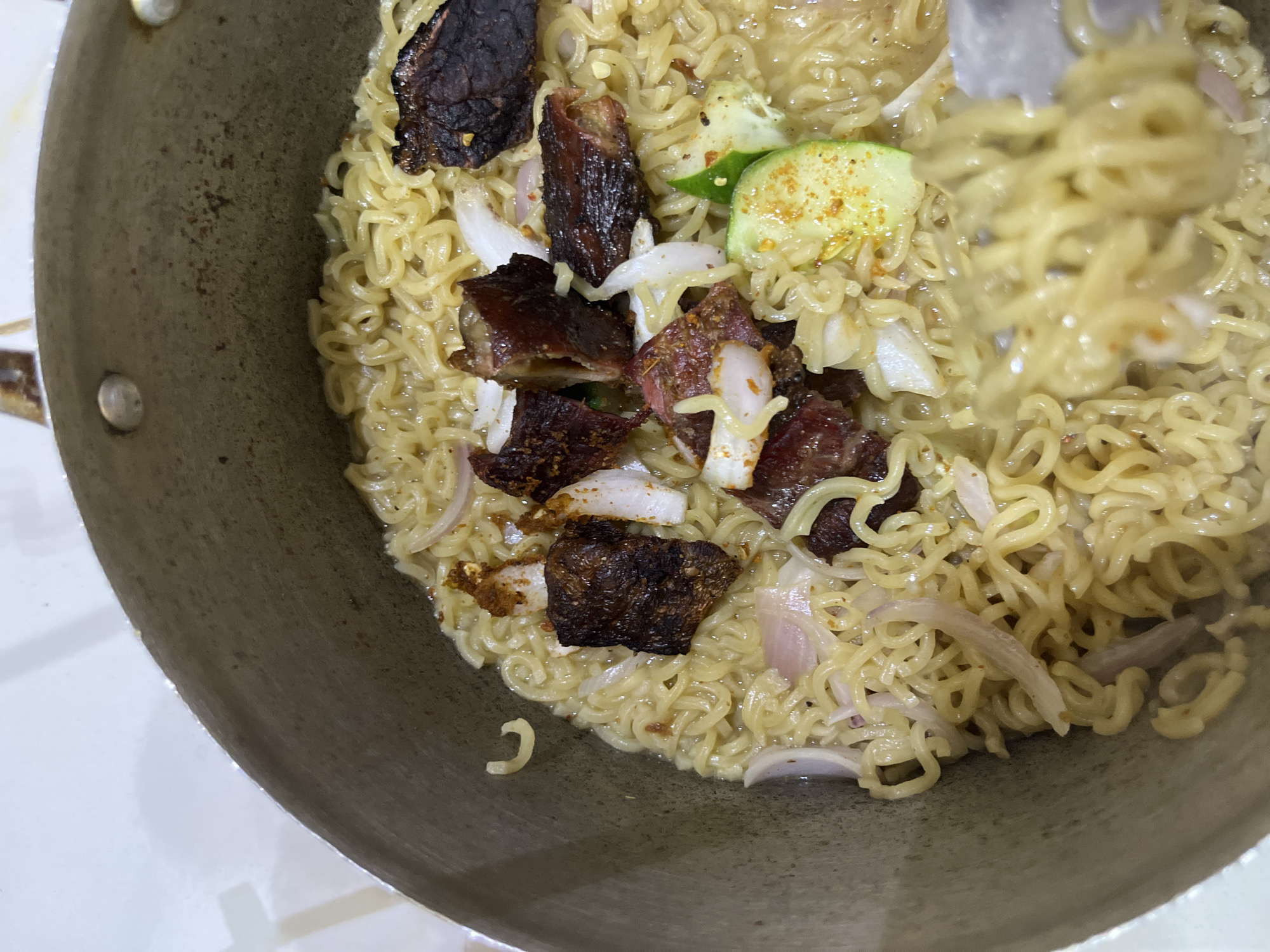 This is a close-up, overhead shot of instant noodles in a pot, likely in Abuja, Nigeria. The noodles are a pale yellow and appear to be cooked in a broth. They are topped with pieces of dark, cooked meat, thinly sliced red onion, and some green vegetable, possibly cucumber, sprinkled with a yellow spice. The pot is made of metal and has a worn appearance. The background is out of focus but appears to be a light-colored surface, possibly a countertop or table.