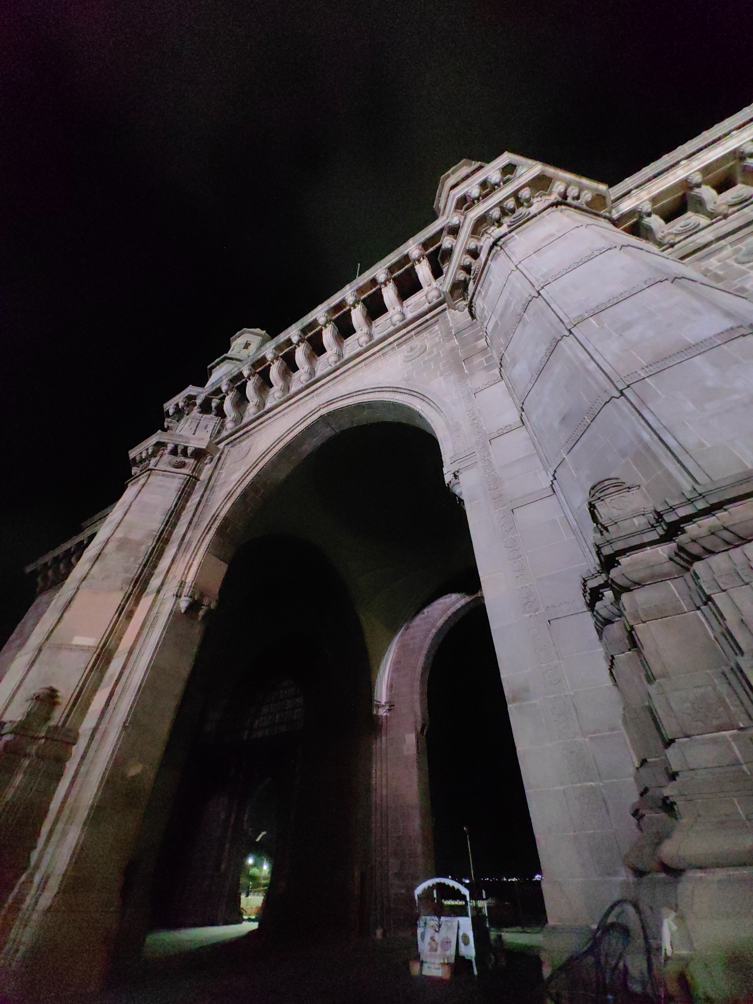 The image shows a low-angle, nighttime shot of the Gateway of India, a prominent monument located in Apollo Bunder, Mumbai, India. The monument, constructed of basalt, is illuminated, revealing its intricate architectural details, including arches, decorative carvings, and a crenellated parapet. The sky above is dark, with some subtle hints of distant lights or possibly colored lighting reflecting from the monument or surrounding areas. In the foreground, beneath the main arch, the scene is darker, with the entrance to the monument casting deep shadows. A small cart or stall with some signage is visible on the right side, and a streetlamp is faintly visible in the distance through the archway, illuminating a pathway. The overall impression is one of grandeur and historical significance, captured at night.