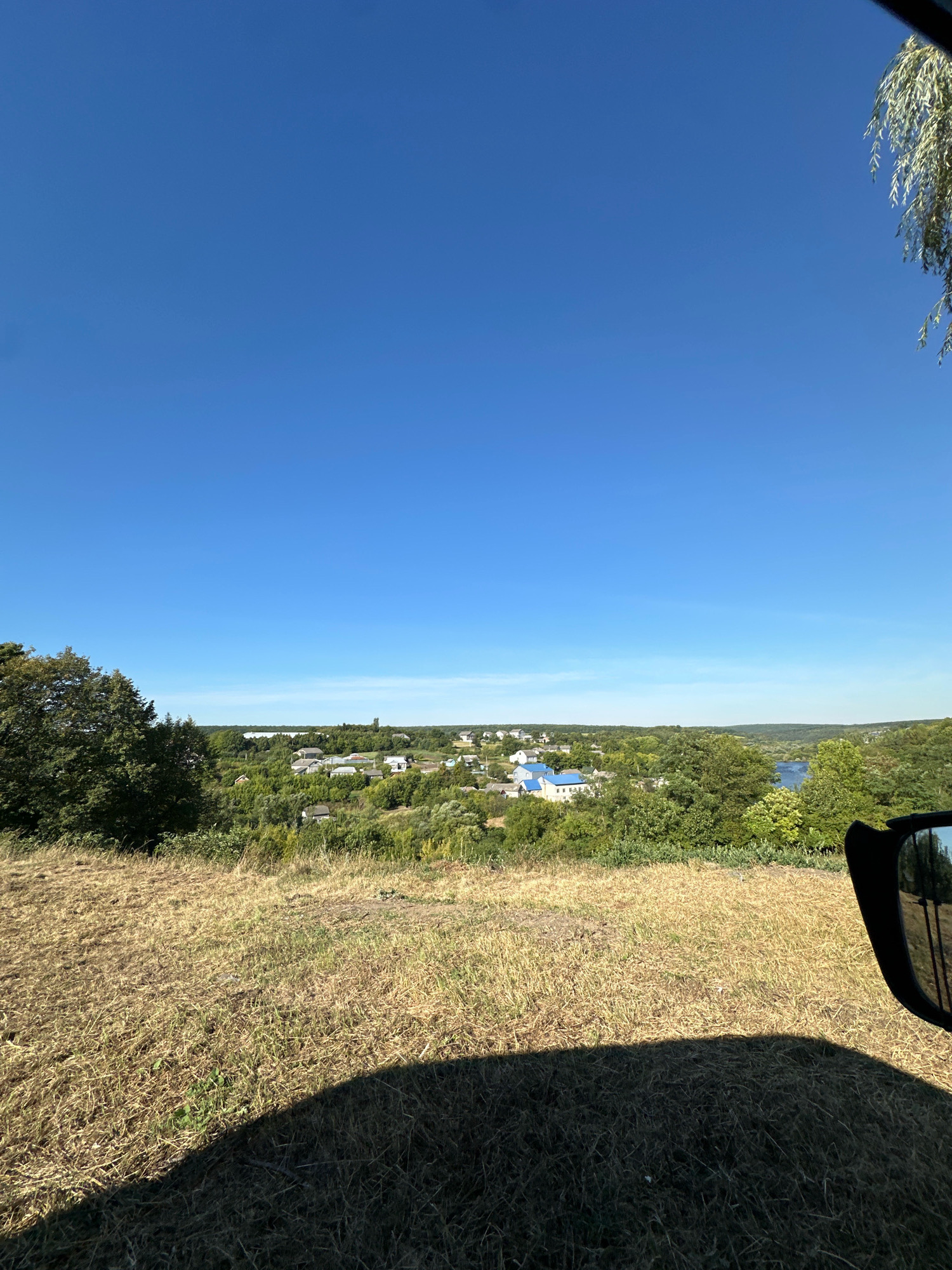 The image depicts a panoramic view of a rural landscape under a clear blue sky. In the foreground, a dry, grassy hillside casts a shadow from an unseen object, likely a vehicle. A side-view mirror is visible on the right edge, reflecting trees and a distant structure. The middle ground showcases a village with numerous houses, many featuring white walls and blue roofs, nestled among lush green trees. A body of water, possibly a lake or river, is visible in the distance. The horizon is lined with rolling hills and more greenery. The overall impression is that of a serene, sunlit day in a Ukrainian countryside setting. The scene is devoid of any people or animals.