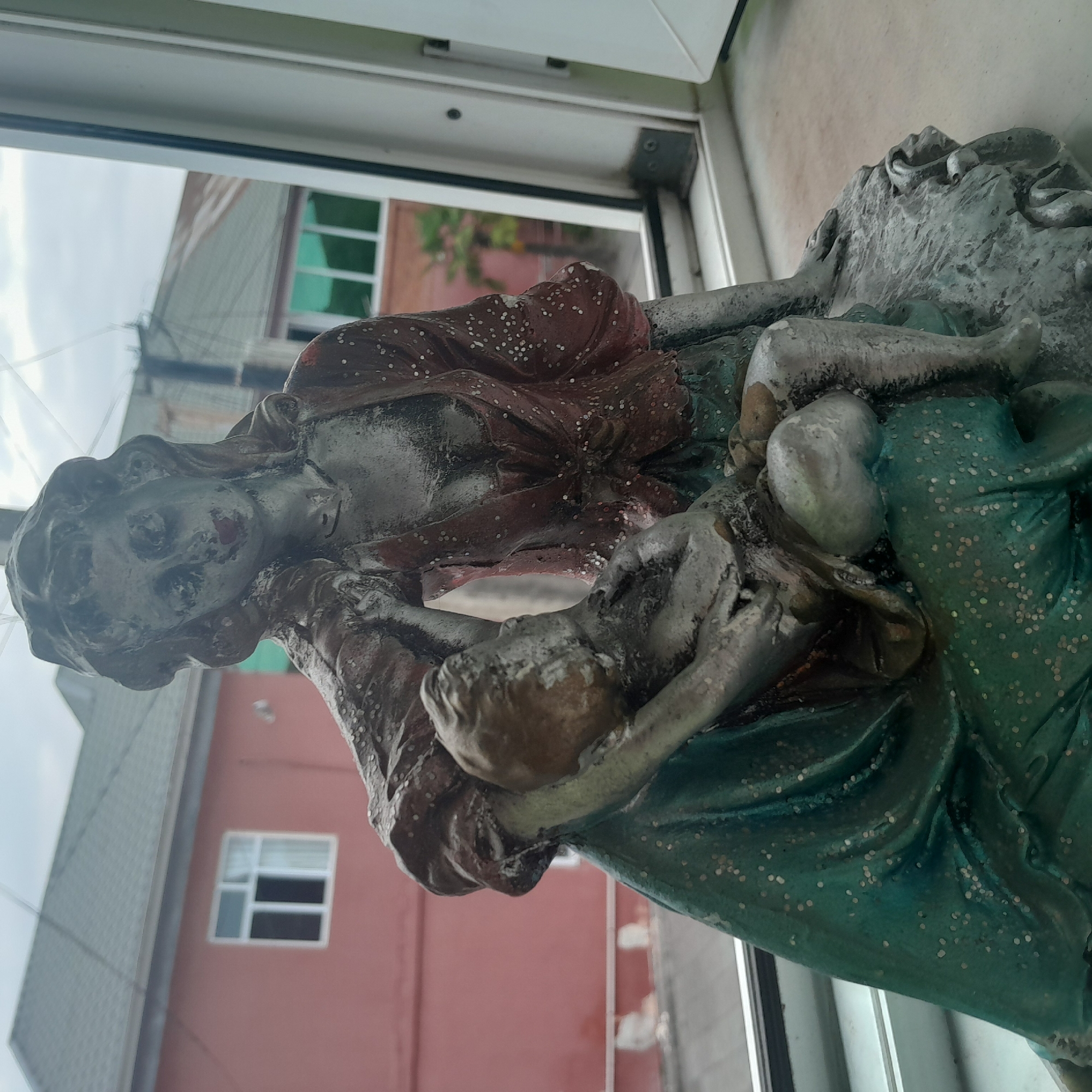The image shows a statue of a woman holding a child. The statue is made of what appears to be aged metal or resin with a patina. The woman has long, wavy hair and is wearing a red garment with white polka dots. She is leaning back, with her hand resting on her head. The child is cradled in her lap, also appearing aged and somewhat indistinct. The statue is placed on a windowsill, with a view of buildings and the sky through a window. The buildings in the background are a faded pink and a lighter color, with green-paned windows. A tree is visible to the right of the pink building. The sky is overcast. The scene is likely in Astara, Azerbaijan, based on the architectural style and the presence of what could be a palm tree.