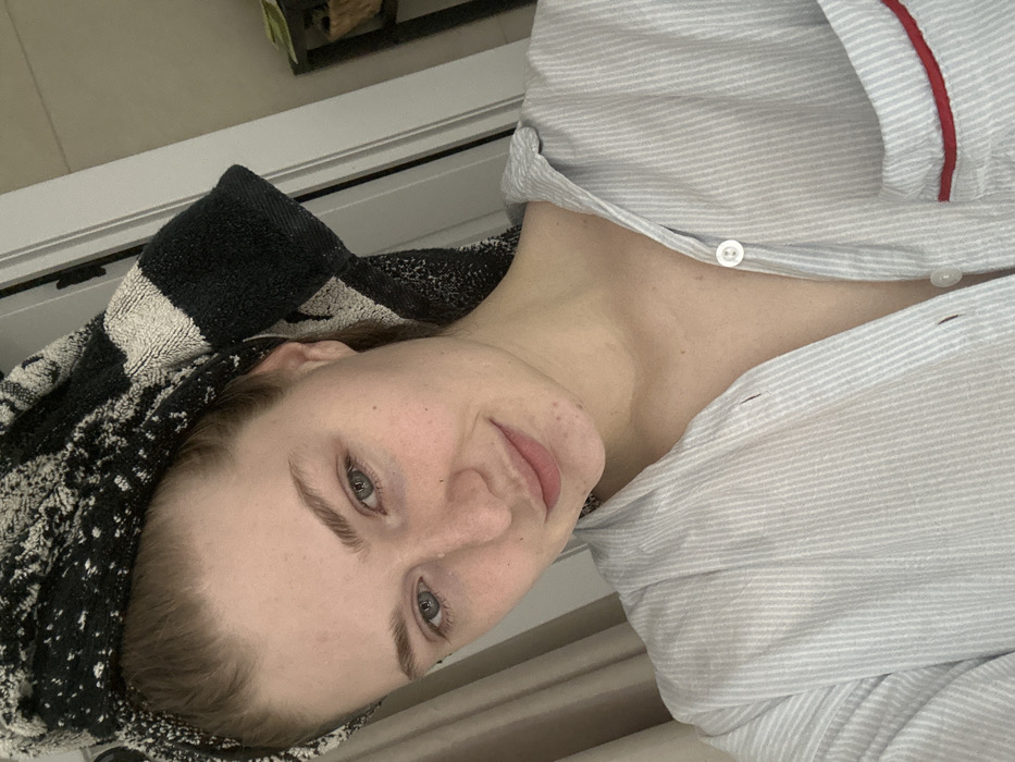A woman with light skin and hair is lying down. She has a towel wrapped around her head and is wearing a striped pajama top with red piping. She looks at the camera with a slight smile. Her eyes are blue-grey. A window frame can be seen in the background, with a glimpse of a potted plant. The image is rotated, giving a tilted perspective.