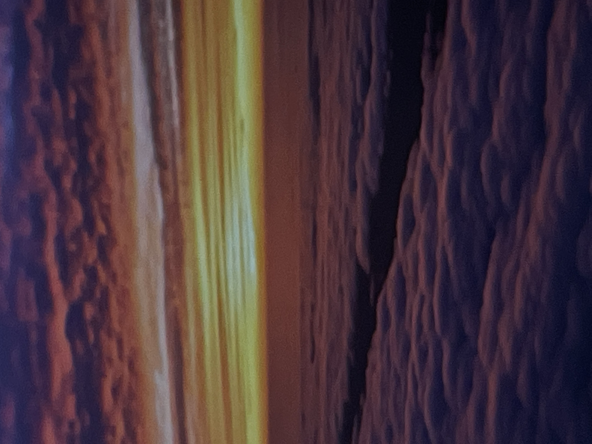 The image depicts a twilight sky over a sea of clouds, likely taken from an airplane or high vantage point. The sky exhibits a gradient of colors, transitioning from deep purple and red hues near the top to vibrant yellow and orange at the horizon. Wispy clouds are scattered across the upper portion of the image, catching the fading sunlight. Below the horizon, a dense layer of fluffy clouds fills the lower half of the frame, with hints of purple and pink tones due to the ambient light. There is no discernible city, person, or specific object present in the scene. The location in Denpasar, Indonesia is not identifiable from the visual information provided.