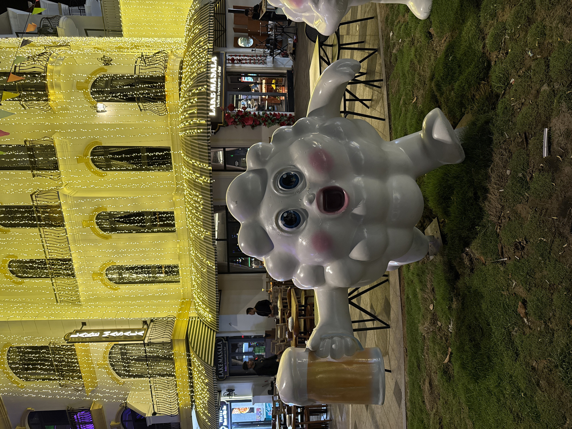 The image captures a vibrant night scene in Phú Quốc, Vietnam, illuminated by numerous warm-toned string lights. The central subject is a large, white, cartoonish figure resembling a cloud or fluffy animal, with wide blue eyes, pink cheeks, and an open mouth suggesting surprise or excitement. This whimsical character holds a large mug filled with a frothy, beer-like beverage.
Behind the character is a multi-story building, likely a commercial complex, heavily adorned with bright yellow string lights that drape across its facade and balconies. A restaurant, prominently identified by signs reading "LAKE MASALA Indian Restaurant & Bar," is visible on the ground floor. It features a striped awning and outdoor seating areas with tables and chairs. A few indistinct figures of people can be seen sitting at tables or standing near the entrance of the restaurant, indicating activity. One person is clearly visible standing near the entrance of the restaurant, and another can be seen sitting inside. Decorative red flowers are arranged near the restaurant's entrance, and colorful flags (bunting) hang across the building's upper levels. In the foreground and to the side, a patch of green grass and dirt is visible, contrasting with the paved area where the character stands. The overall atmosphere is festive and inviting, characteristic of an evening entertainment or dining district.