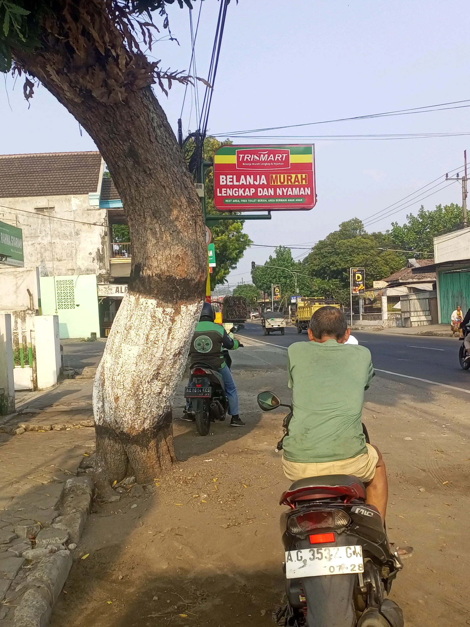The image depicts a street scene in Gurah, Indonesia, on a bright, clear day. In the foreground, a person is seated on a motorcycle, viewed from behind. The motorcycle has a license plate that reads "AG 3532 GW 07-28". To the left of the motorcycle, a large tree with a thick, textured trunk dominates the frame. The lower part of the tree trunk has been painted with white and dark brown bands. A red and green billboard with the text "TRISMART BELANJA MURAH LENGKAP DAN NYAMAN" is visible behind the tree, suspended from poles.
The background shows a paved road with other vehicles, including a yellow truck and other motorcycles. Buildings line the street, some with tiled roofs and what appear to be shops or businesses. Trees and greenery are also present, suggesting a semi-urban or suburban environment. The overall impression is of a typical day in a bustling Indonesian town, with everyday activities like motorcycling and commerce taking place.