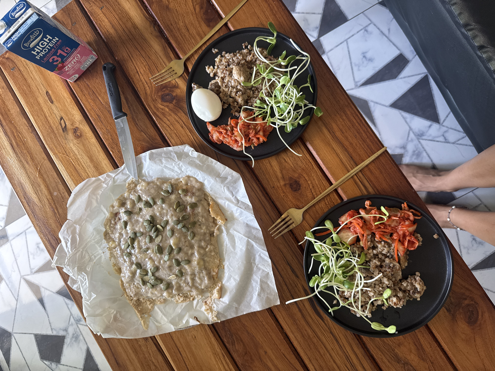 The image shows a wooden table with two black plates of food, a piece of food on parchment paper, a carton of Benefitt high protein lactose-free milk, and a knife. One plate contains a boiled egg, cooked grains, kimchi, and sprouts. The other plate contains cooked grains, kimchi, and sprouts. The food on parchment paper is spread with a beige substance and topped with pumpkin seeds. There are two golden forks on the table. Parts of two human legs are visible at the right edge of the image, and a tiled floor is visible beneath the table. The location in the city of Moo 4, Thailand is not determinable from the image.
