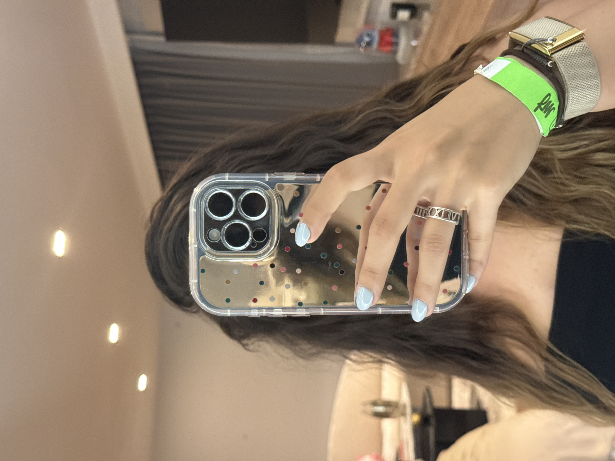 A woman holds a smartphone up to a mirror, capturing a selfie. The phone is encased in a clear case adorned with multicolored polka dots. The woman's fingers are painted a light blue. She wears a silver ring inscribed with Roman numerals on one finger. On her wrist, she wears a watch with a brown leather band and a woven beige strap, along with a bright green wristband featuring black text. The woman's dark, wavy hair is visible behind the phone. The background includes a wall, lighting fixtures, and a glimpse of an interior with various items and fixtures. The location is implied but not definitively confirmed to be in San Bernardino Tlaxcalancingo, Mexico.
