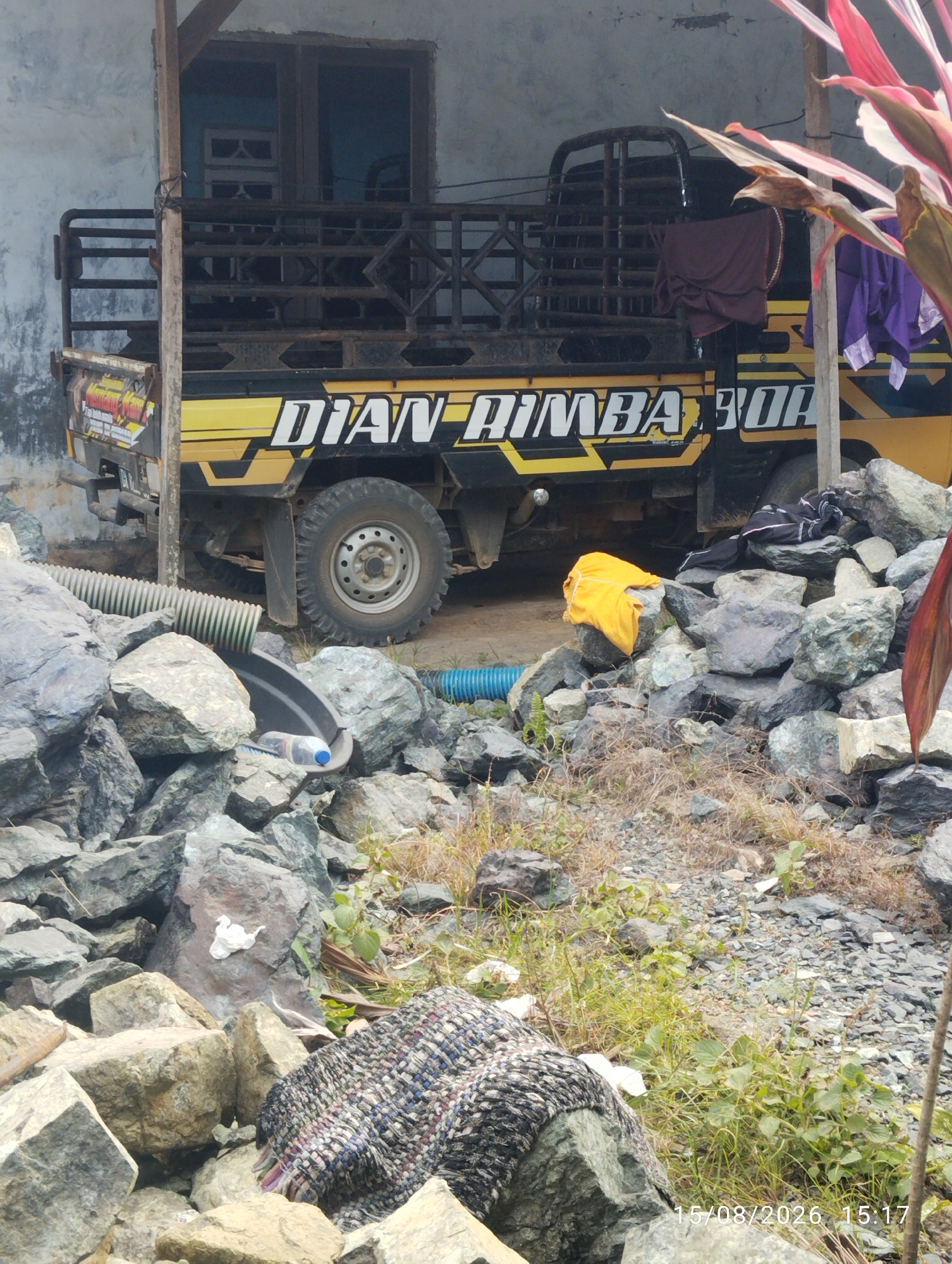 The image depicts an outdoor scene at what appears to be a residential or storage area. In the background, a yellow pickup truck with black accents and the words "DIAN RIMBA BO" visible on its side is parked under a simple wooden shelter. The truck has a metal cage-like bed. To the right of the truck, there are two pieces of clothing hanging, one dark brown and one purple.
The foreground is dominated by a large pile of gray and tan rocks of various sizes. Among the rocks, there is a black plastic drainage pipe with a ribbed surface, leading to a curved black trough where a clear plastic bottle with a blue cap is placed. A blue corrugated pipe is also visible in the middle ground. A yellow piece of fabric is draped over a rock. In the lower right quadrant of the image, a woven fabric or rug with a patterned design is partially visible, lying on the rocks. The ground is covered with gravel and dry vegetation. In the lower right corner of the image, a timestamp reads "15/08/2026 15:17". The overall impression is of a somewhat unkempt or work-in-progress outdoor space. The location is identified as Tegal Rejo, Indonesia.