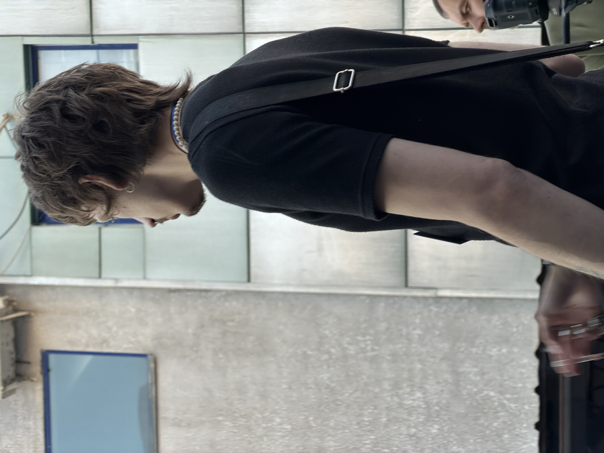 A medium shot, oriented vertically, shows a person with light brown, wavy hair and a black shirt, facing left. They are wearing a beaded necklace and carrying a camera with a strap over their shoulder. Their right arm is partially visible, and they are wearing multiple rings.
The background consists of a gray wall with rectangular panels. A blue-framed window is visible on the left side. Another person is partially visible in the upper right corner, holding a camera. The photograph was taken in Bishkek City, Kyrgyzstan.