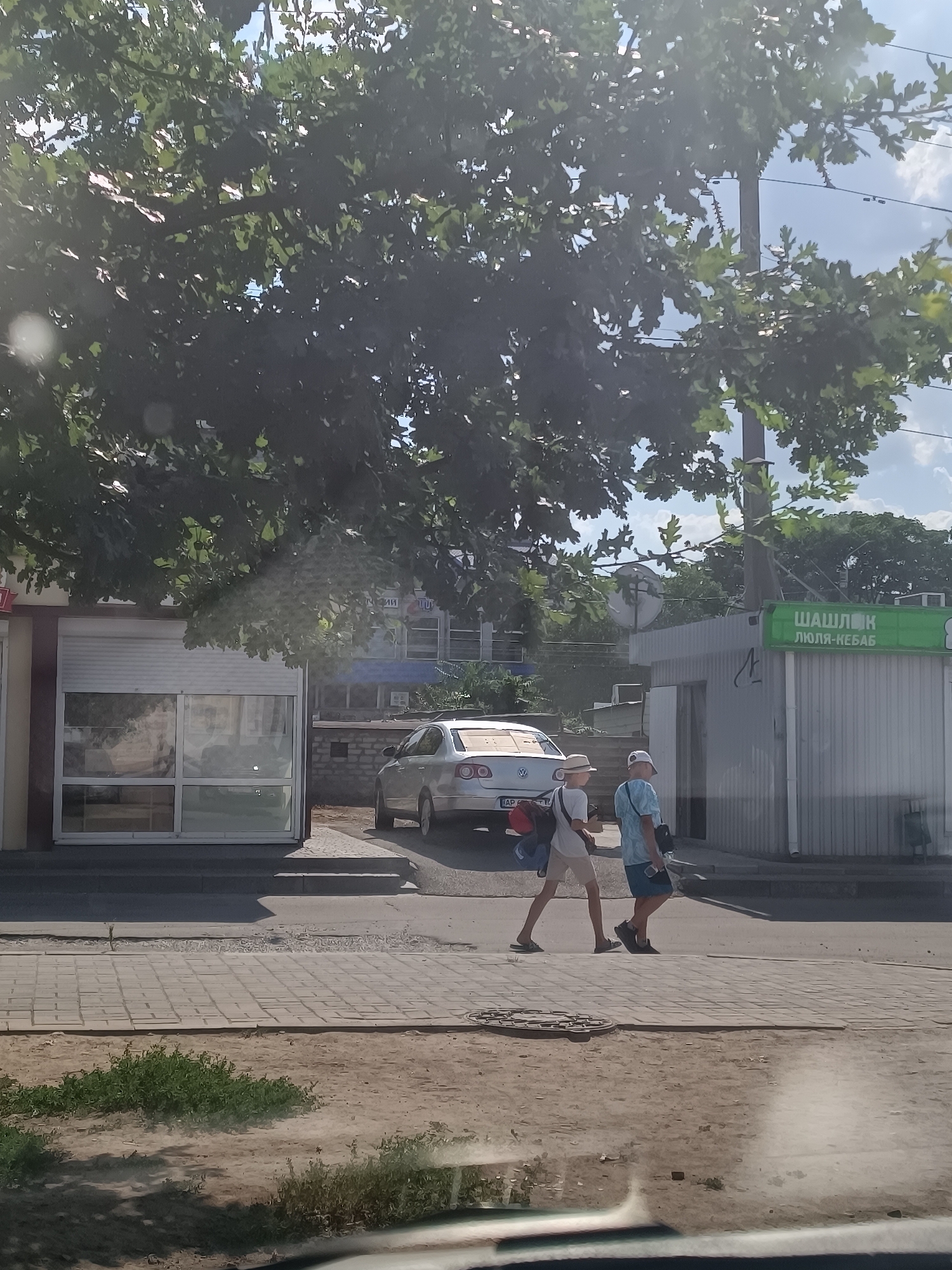 The image shows a street scene in Zaporizhzhia, Ukraine, on a sunny day. Two young people, a male and a female, are walking on a paved sidewalk. The male is wearing a white hat, a white t-shirt, and light-colored shorts, carrying a backpack over his right shoulder. The female is wearing a light blue t-shirt, dark blue shorts, and sunglasses, carrying a bag strap over her left shoulder. They appear to be walking away from the camera.
In the background, there is a silver car parked on the street, possibly a Volkswagen. Adjacent to the car is a small building with a white shuttered window and glass panels. Further to the right, there is a light-colored, corrugated metal structure with a green sign that has Cyrillic lettering, reading "ШАШЛЫК" (Shashlyk) and "ЛЮЛЯ-КЕБАБ" (Lyulya-kebab), indicating a food establishment. Behind these structures, there are trees with lush green foliage and a cloudy sky. The overall impression is a casual outdoor environment in an urban setting.