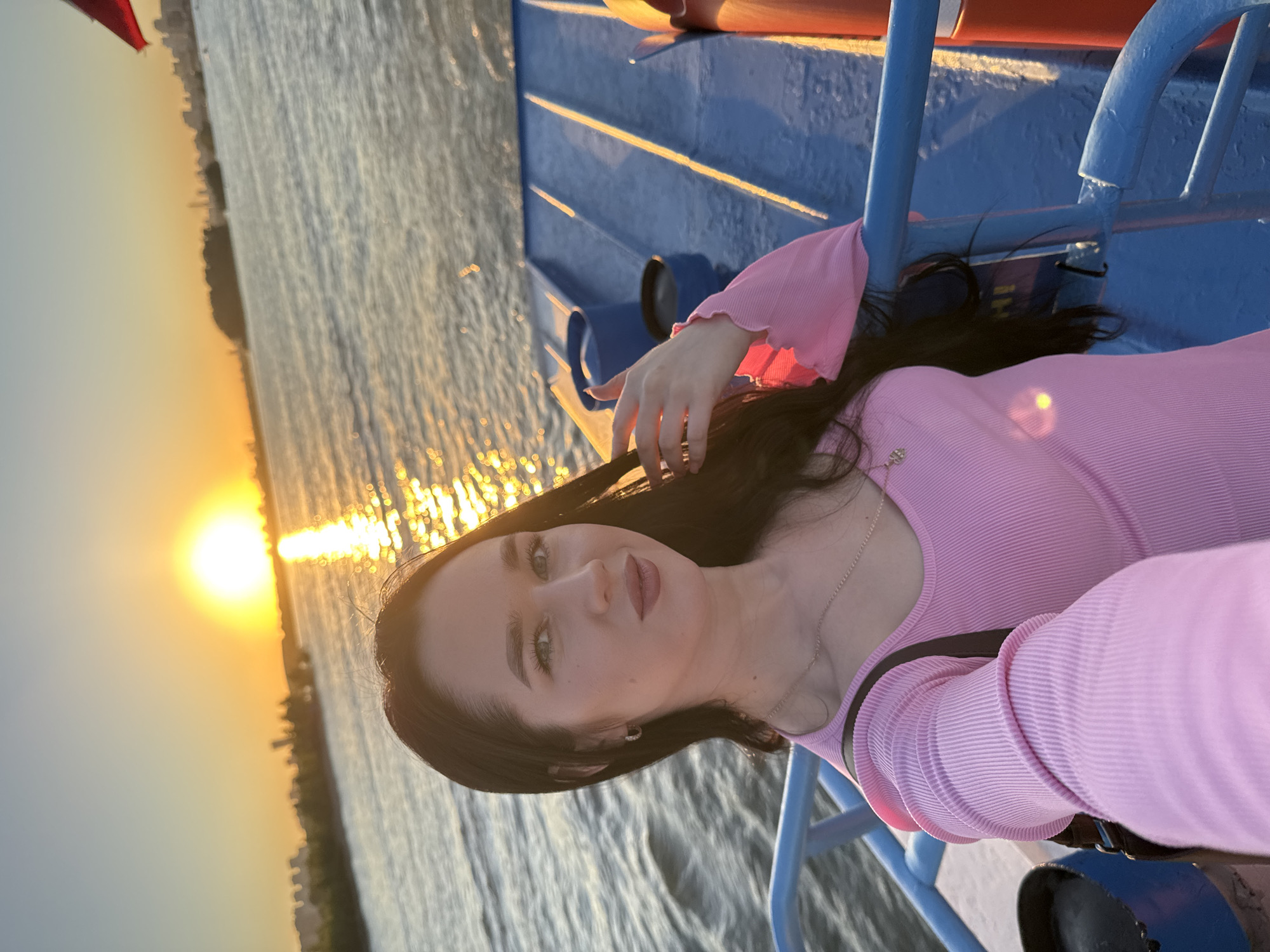 A young woman with long dark hair takes a selfie on a boat at sunset. She is wearing a pink ribbed long-sleeved top and a necklace. The sun is setting over the water, casting a golden glow. In the distance, a cityscape is visible, likely the Novosibirsk skyline. The boat has blue railing and seating. The water appears choppy and reflects the sunset.
