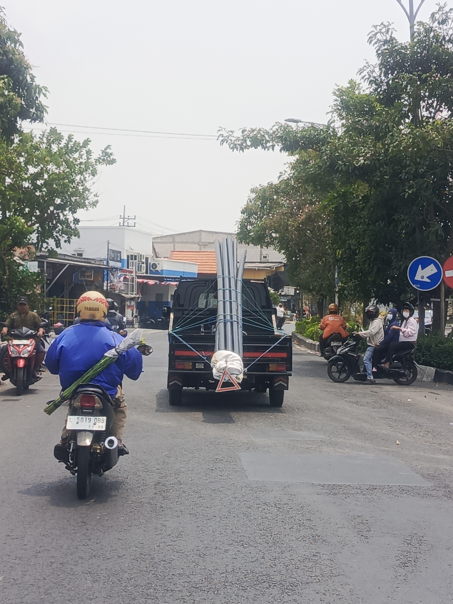 In the city of Surabaya, Indonesia, on a cloudy day, a motorcycle rider wearing a blue jacket and a yellow helmet with "YAMAHA" printed on it is seen from behind. The rider is carrying a bundle of long green reeds tied together, extending from their side to the rear of the motorcycle. Another motorcycle with a red body is to the left of the rider. In front of the motorcycle rider is a black truck carrying a load of long metal pipes, secured with ropes. The truck has a red and white triangular marker at the rear. Further ahead on the right side of the road, two motorcycles are visible. One is black, carrying three passengers wearing helmets, and the other is orange, carrying a single rider. A circular blue sign with a white arrow indicating a left turn and a red circular sign are present on the right side of the road. The road surface appears to be asphalt with some patched areas. Trees line the right side of the road, and buildings with various signage are visible in the background.