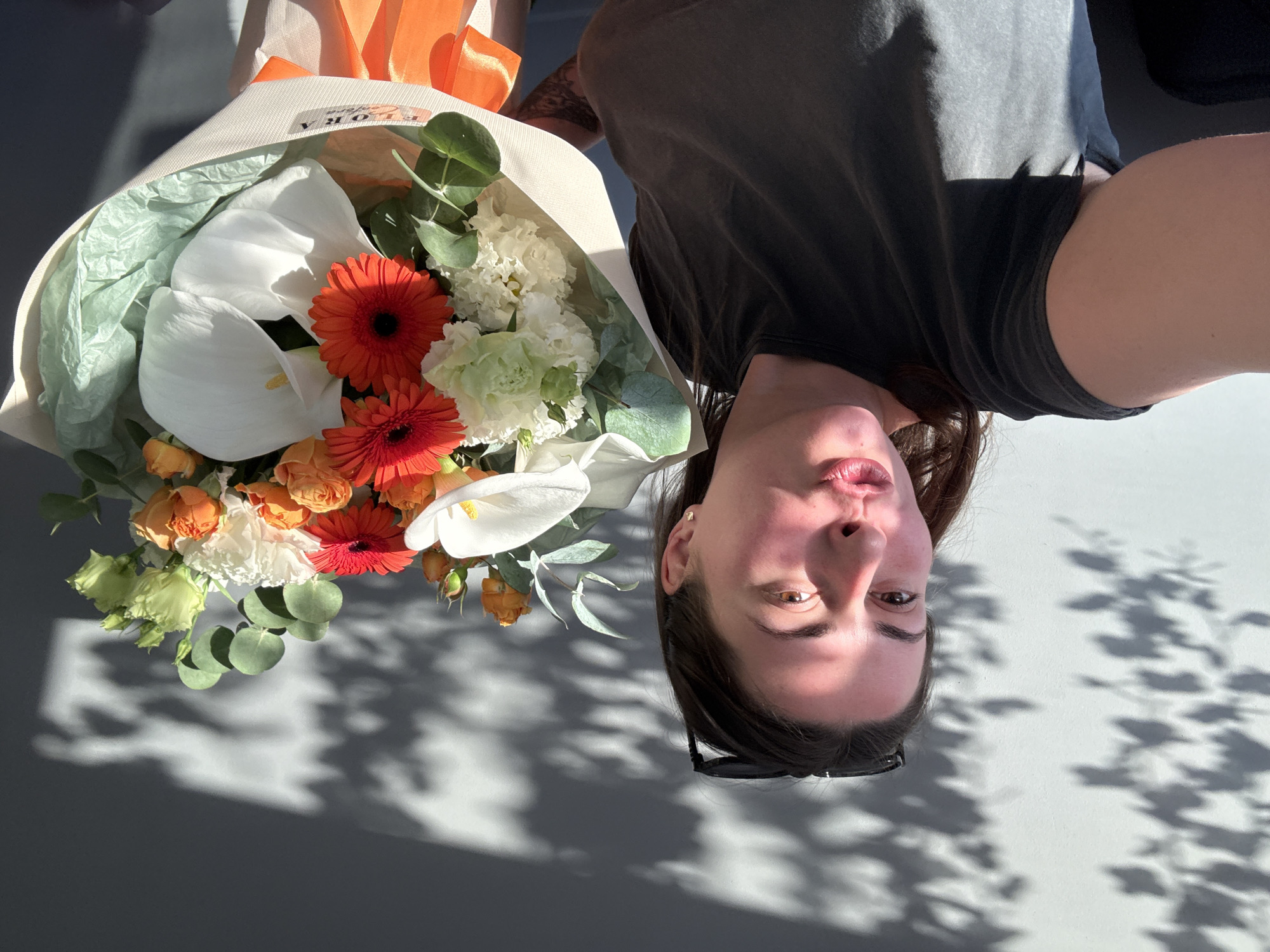In this medium shot, a woman with dark hair is taking a selfie in front of a white wall with dappled shadows cast by sunlight. She is wearing a dark grey t-shirt and has sunglasses perched on her head. Her lips are puckered, and she is holding a bouquet of flowers that includes white calla lilies, red gerbera daisies, orange roses, and greenery. The bouquet is wrapped in cream paper and tied with an orange ribbon. The lighting suggests it is daytime, and the shadows indicate the presence of foliage outside.