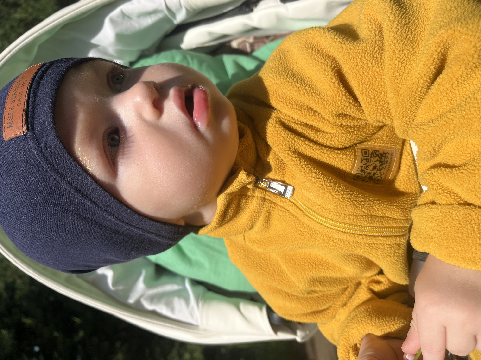 A young child, appearing to be a toddler, is depicted in a medium shot, seated outdoors in a stroller in Сосново, Russia. The child wears a dark blue knit beanie with a brown rectangular label featuring text "MSEK SEANS SINCE 1993", and a bright yellow fleece jacket with a full-length zipper and a silver-toned zipper pull. A black and white QR code label with partially visible text "mamyasam" is affixed to the right sleeve of the jacket. The child has fair skin, light-colored hair, and light eyes, looking towards the right of the frame with a slightly open mouth. The child's right hand, visible in the lower left, holds a small green object. The background consists of blurred dark green foliage and the light-colored interior lining of the stroller. The scene is illuminated by direct sunlight.