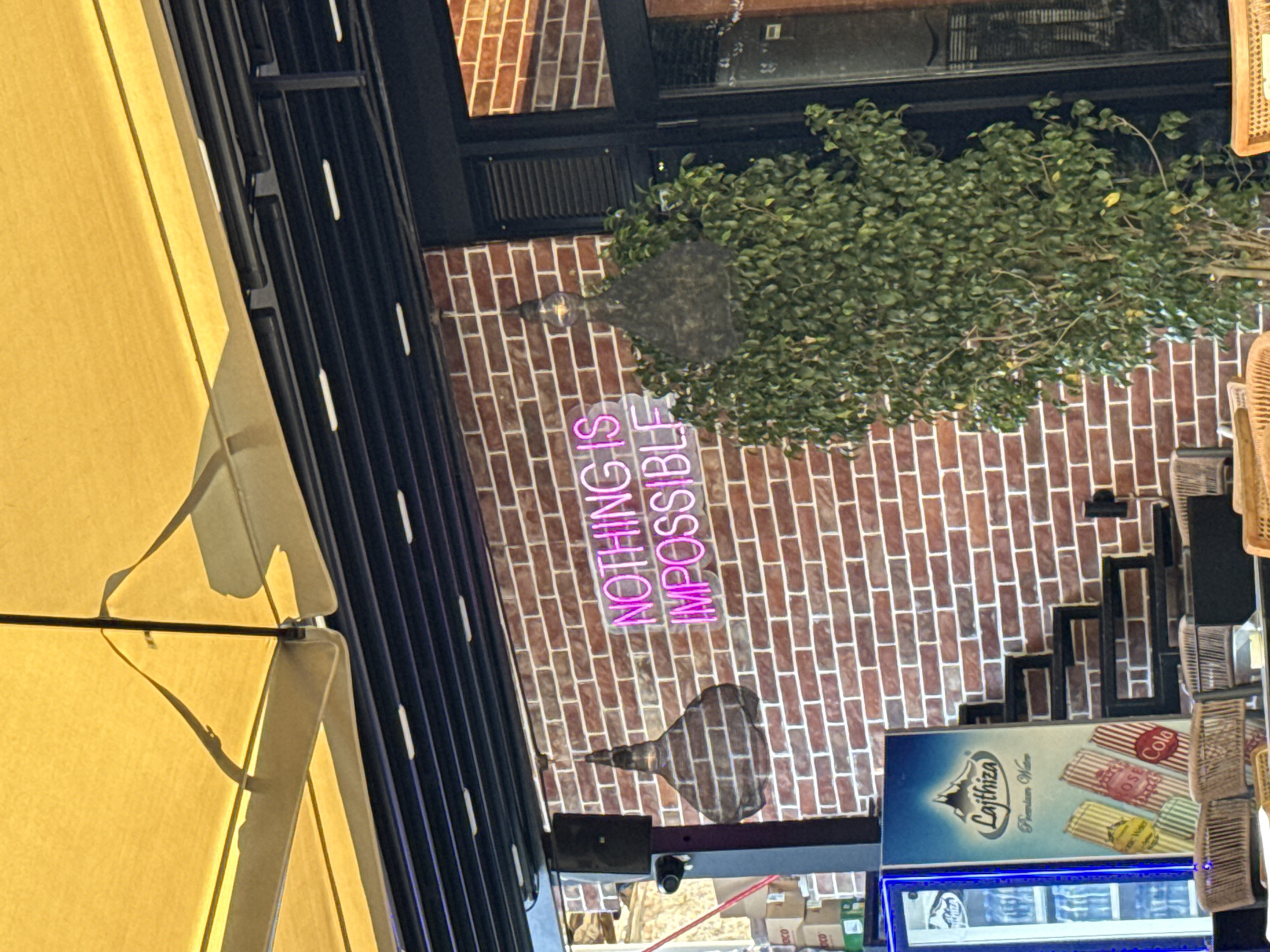 The image shows a brick wall with a neon sign that reads "NOTHING IS IMPOSSIBLE". To the left of the sign, there is a black speaker and a security camera mounted on a black pole. Below the speaker, there is a refrigerator with a label that says "Lajthiza Premium Water". To the right of the sign, there is a tall green plant. In the foreground, there are wicker chairs. The scene appears to be an outdoor seating area of a cafe or restaurant, possibly in Elbasan, Albania, given the presence of the "Lajthiza" brand which is popular in Albania. The lighting suggests it is daytime.