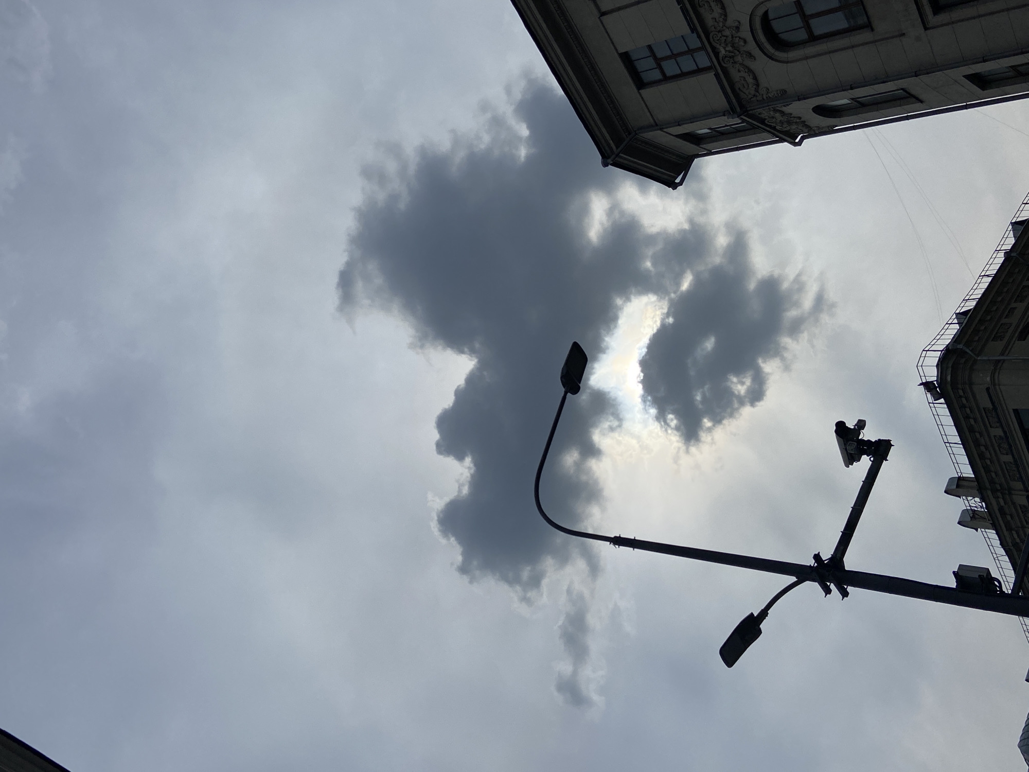 The image, taken from a low angle, depicts a cloudy sky with the silhouettes of streetlights and building facades in Moscow, Russia. The sky is predominantly gray with a large, dark cloud formation in the center. The sunlight is partially obscured by the clouds. Two streetlights are visible: one is a single, curved lamppost, and the other is a complex structure with multiple lights and a camera. Building facades are visible in the upper corners of the frame, showing architectural details and windows. The overall impression is one of an overcast day in an urban environment.