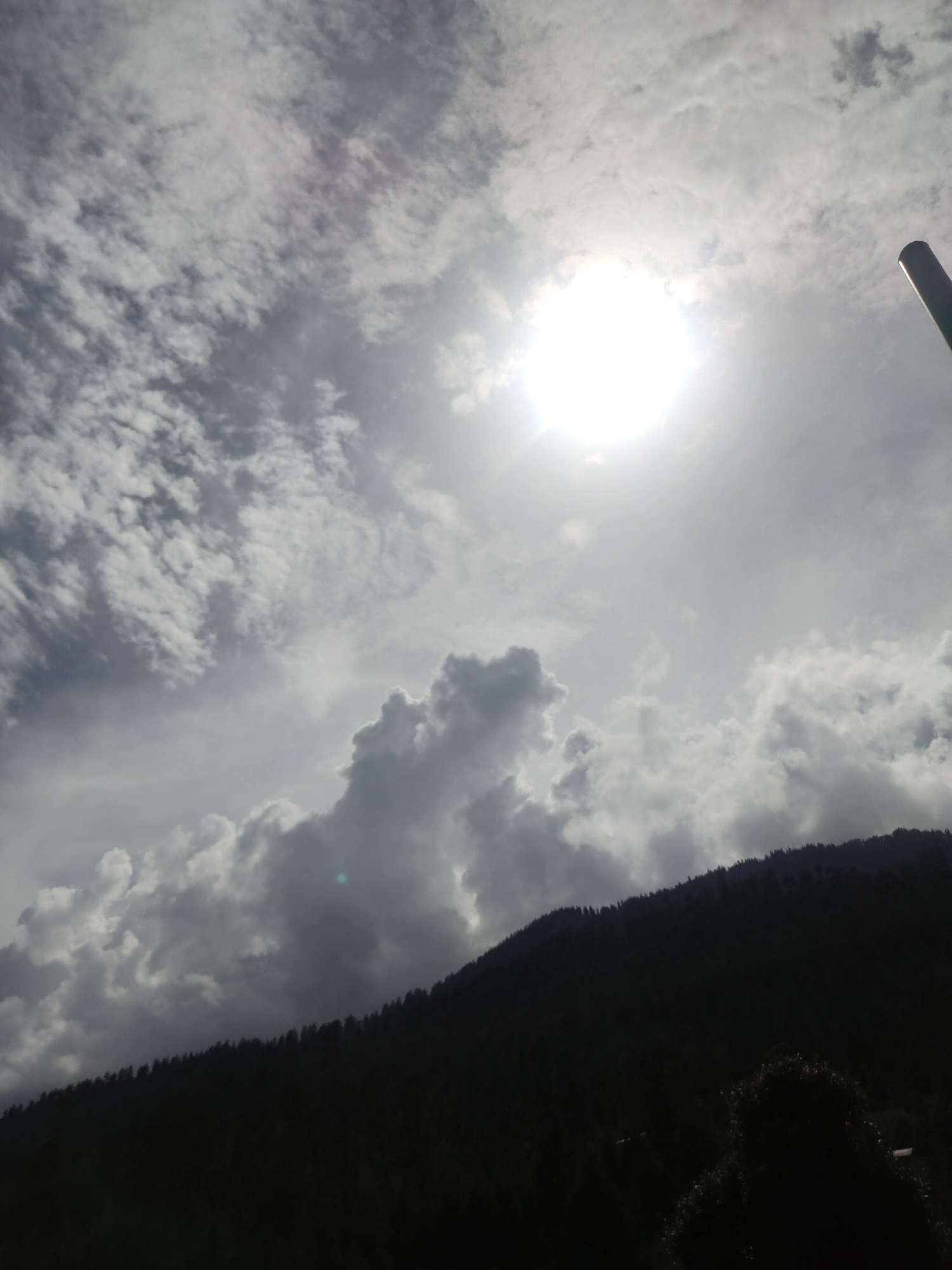 The image shows a bright sun partially obscured by clouds, with a dark, silhouetted mountain range in the foreground. The mountain is covered in trees, forming a dense forest along its slope. The sky is filled with a mix of white and grey clouds, some appearing wispy and others more textured and voluminous. A dark, cylindrical object, possibly a pole or antenna, is visible in the upper right corner of the frame. The overall impression is a dramatic sky over a forested mountain landscape. The location is identified as Manali, India.