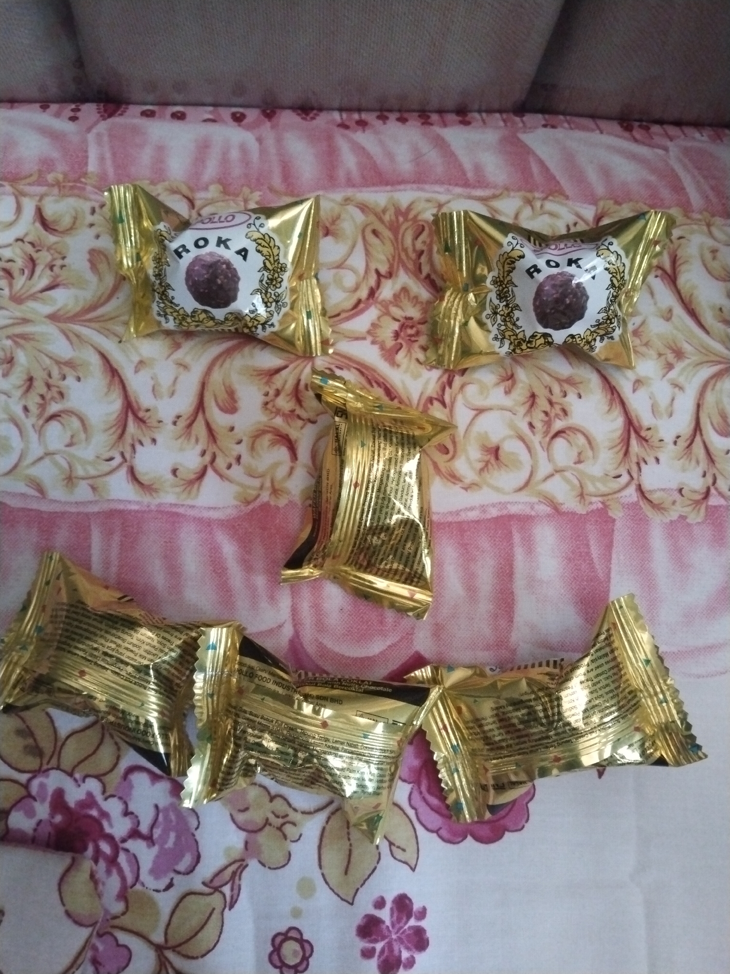 The image shows a flat surface with a patterned bedspread on which are placed six individual, gold-wrapped candies labeled "ROKA." The candies are arranged in a somewhat haphazard pattern, with two near the top, one in the middle, and three along the bottom. The background includes a blurred portion of a sofa or headboard. The information on the wrappers is in Indonesian, suggesting the image was taken in Indonesia. The location in the city of Kutaglumpang, Indonesia is not confirmed by the image.