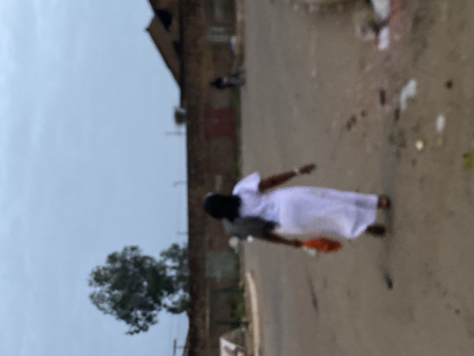 The image, taken in Bukuru, Nigeria, shows a person wearing a white dress and holding an orange object, walking along a street. The environment includes buildings, a tree, and overhead utility lines. The ground appears to be a dirt road. The sky is overcast. The scene suggests an outdoor setting in an urban or semi-urban area.