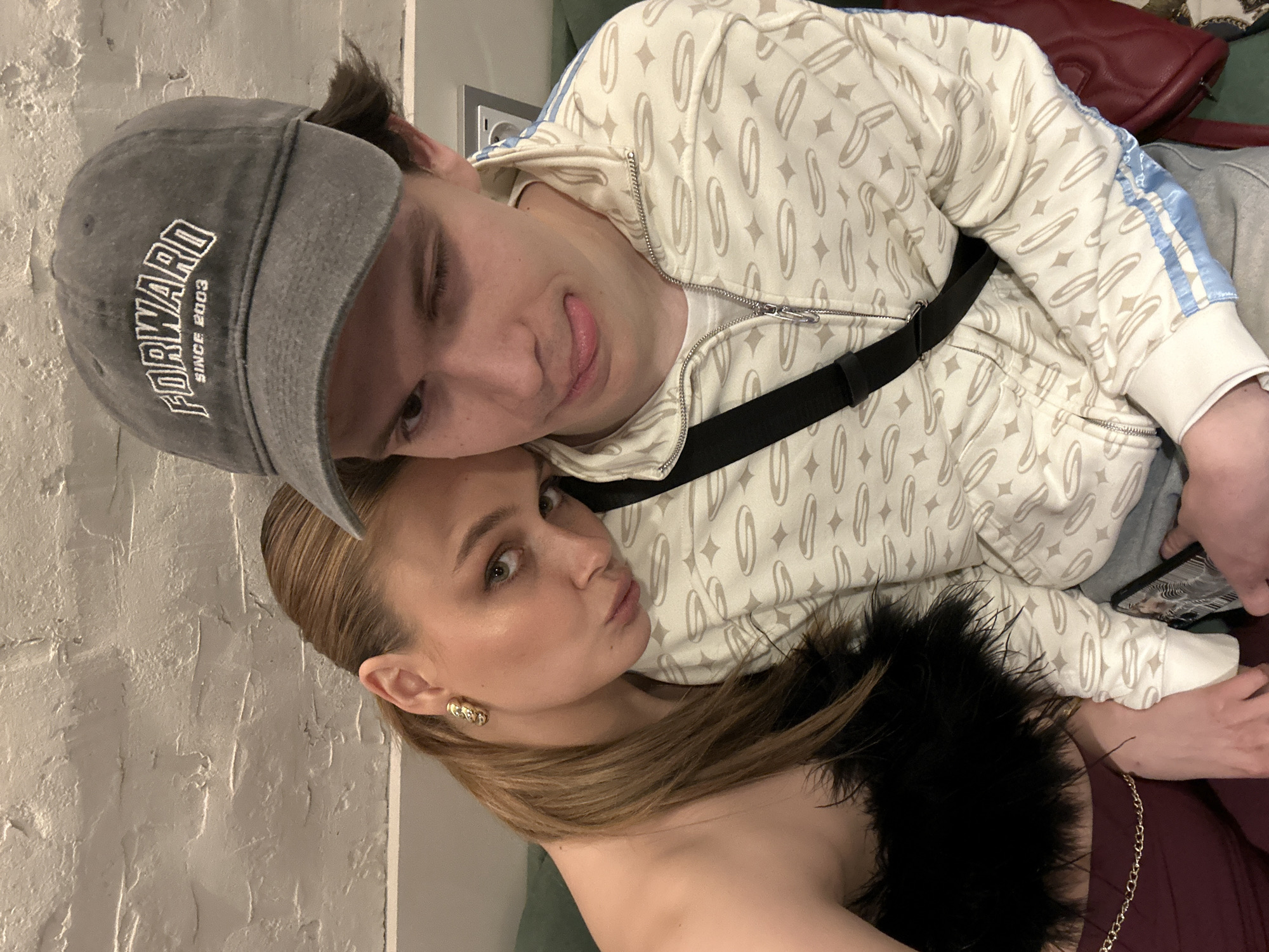 Two individuals are closely positioned, appearing to be posing for a photograph indoors. The person on top is a male wearing a grey baseball cap with the word "FORWARD" embroidered on it, a patterned cream-colored zip-up hoodie with blue stripes on the shoulders, and a black shoulder strap. He is winking and sticking his tongue out. The other person is a female with long brown hair, gold earrings, and a black feathered top. The background includes a textured white wall and a glimpse of what seems like a green sofa or bedding, as well as a burgundy fabric. The location is not determinable from the image.
