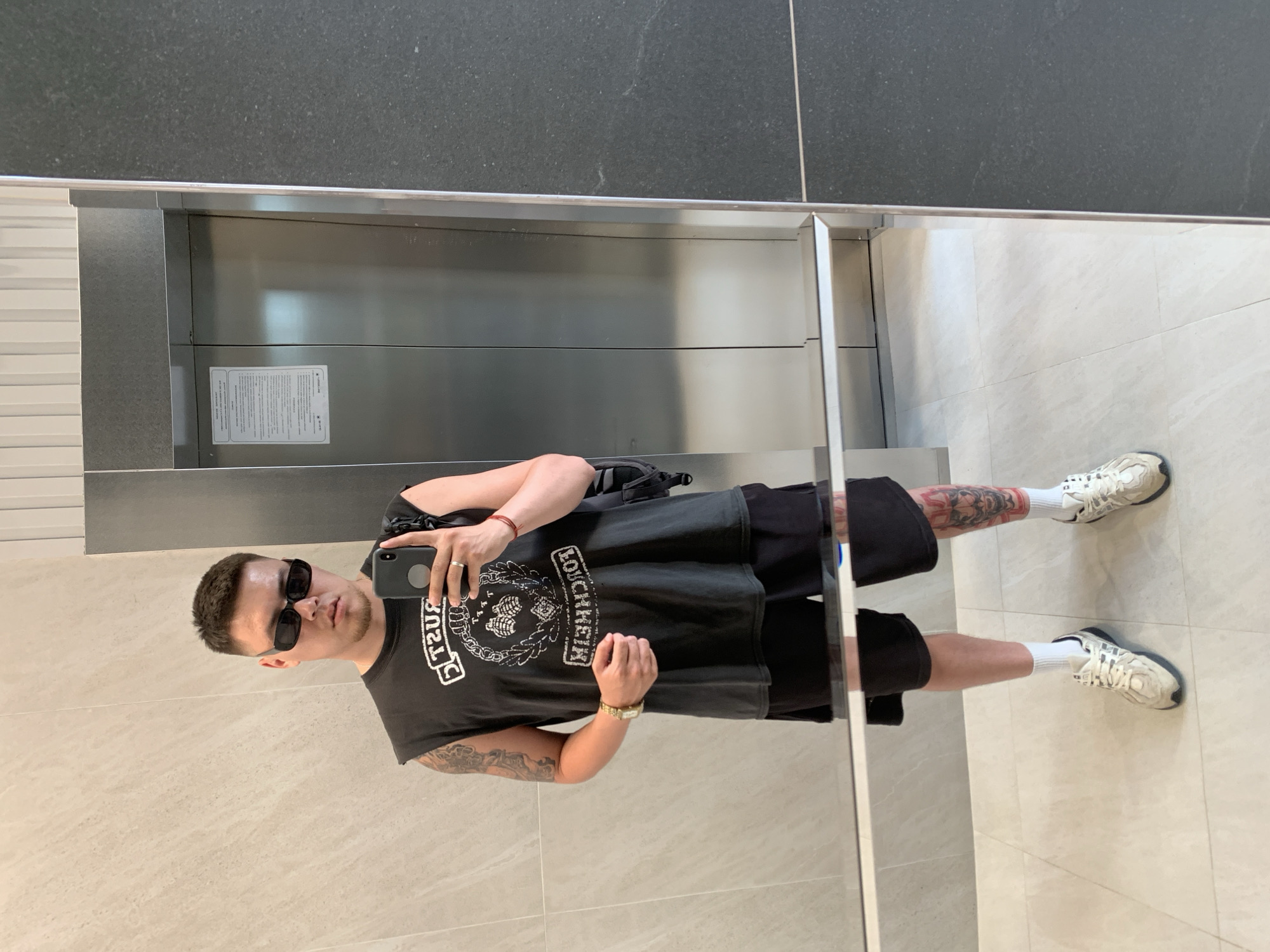 The media file shows a full-length mirror selfie of a young man taken indoors. He is standing in what appears to be a modern building hallway, possibly near an elevator, as suggested by the metallic doors and tiled walls. The man is wearing a black sleeveless t-shirt with a graphic print of a skull and chain, black shorts, white socks, and white athletic shoes. He has sunglasses on and is holding a smartphone in his right hand, taking the picture. A red bracelet is visible on his left wrist, and a ring on his right ring finger. Tattoos are visible on his left arm and right leg. The overall impression is casual and self-aware, with the man presenting himself for the camera. The lighting suggests it is daytime. There is text visible on the t-shirt, although it is partially obscured and reversed due to the mirror. The elevator doors also have a white information sticker on them with text, but it is too small to read.