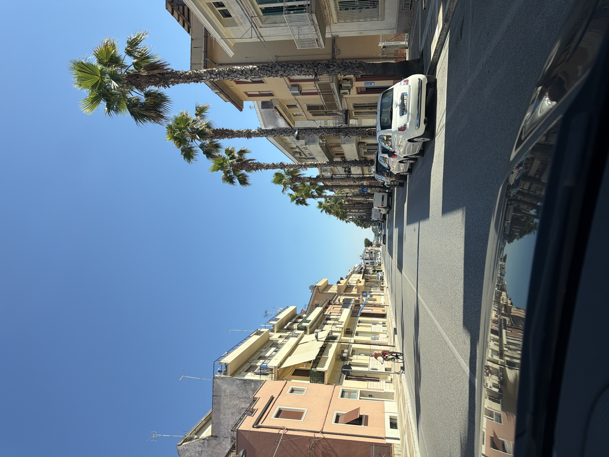 A sunny day scene on a street in Potenza Picena, Italy, likely near the coast, features buildings with balconies and awnings lining both sides of the road. Several tall palm trees stand along the sidewalk, casting shadows on the asphalt. A man is riding a bicycle on the left side of the street. Parked cars are visible along the right side, including a white Fiat 500. The sky is clear and blue. The foreground shows the reflection of the buildings and palm trees on the hood of a dark-colored car.