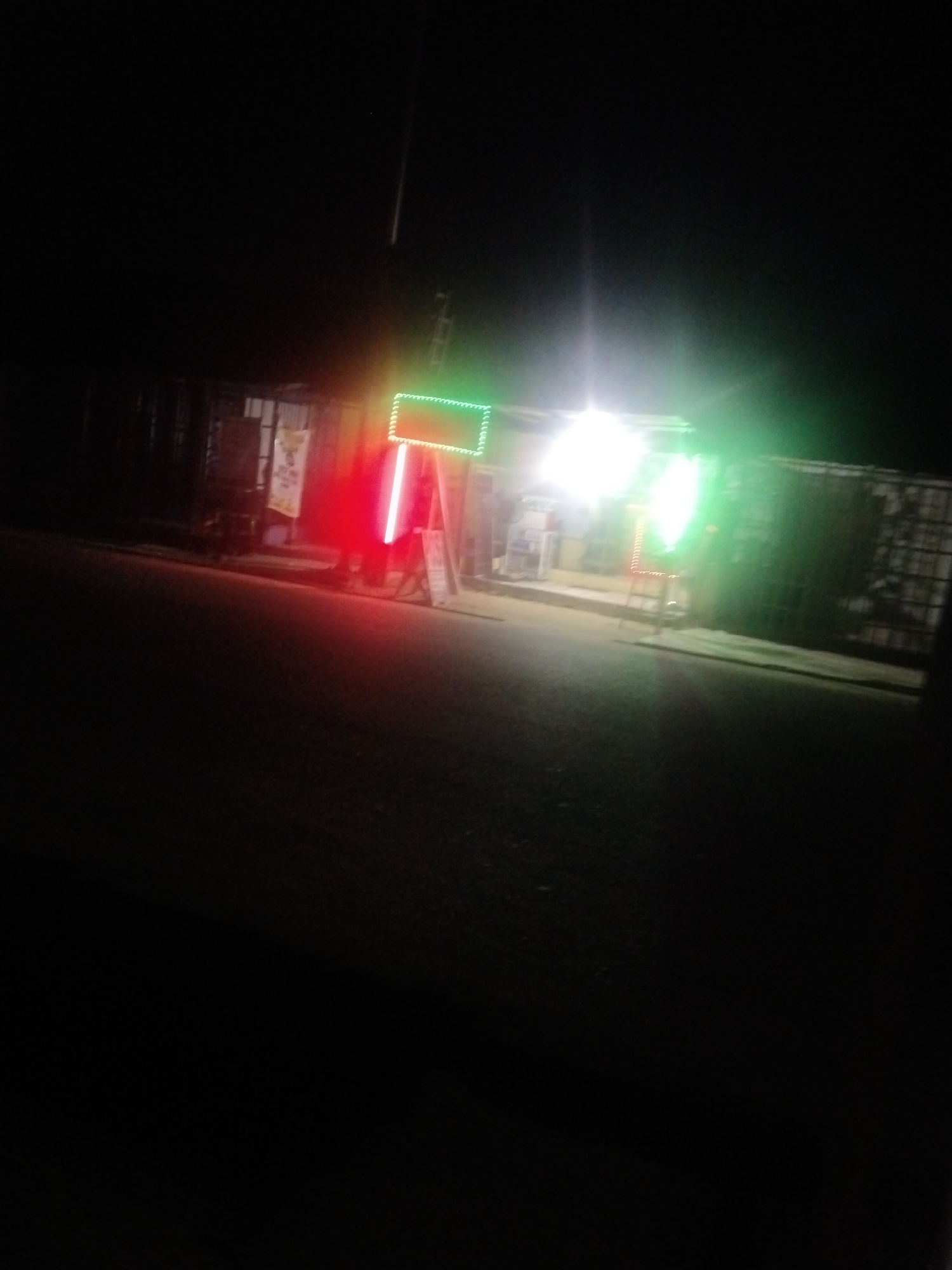 At night on a street in Uyo, Nigeria, a storefront is illuminated by a mix of red and green neon lights and a bright white light. The storefront appears to be a shop or market stall. The street in front of the shop is dark, with the lights from the storefront reflecting on the wet asphalt. The background is obscured by darkness, but some structures and a fence are faintly visible. There are no people clearly visible in the image. The overall scene suggests a lively commercial area at night.