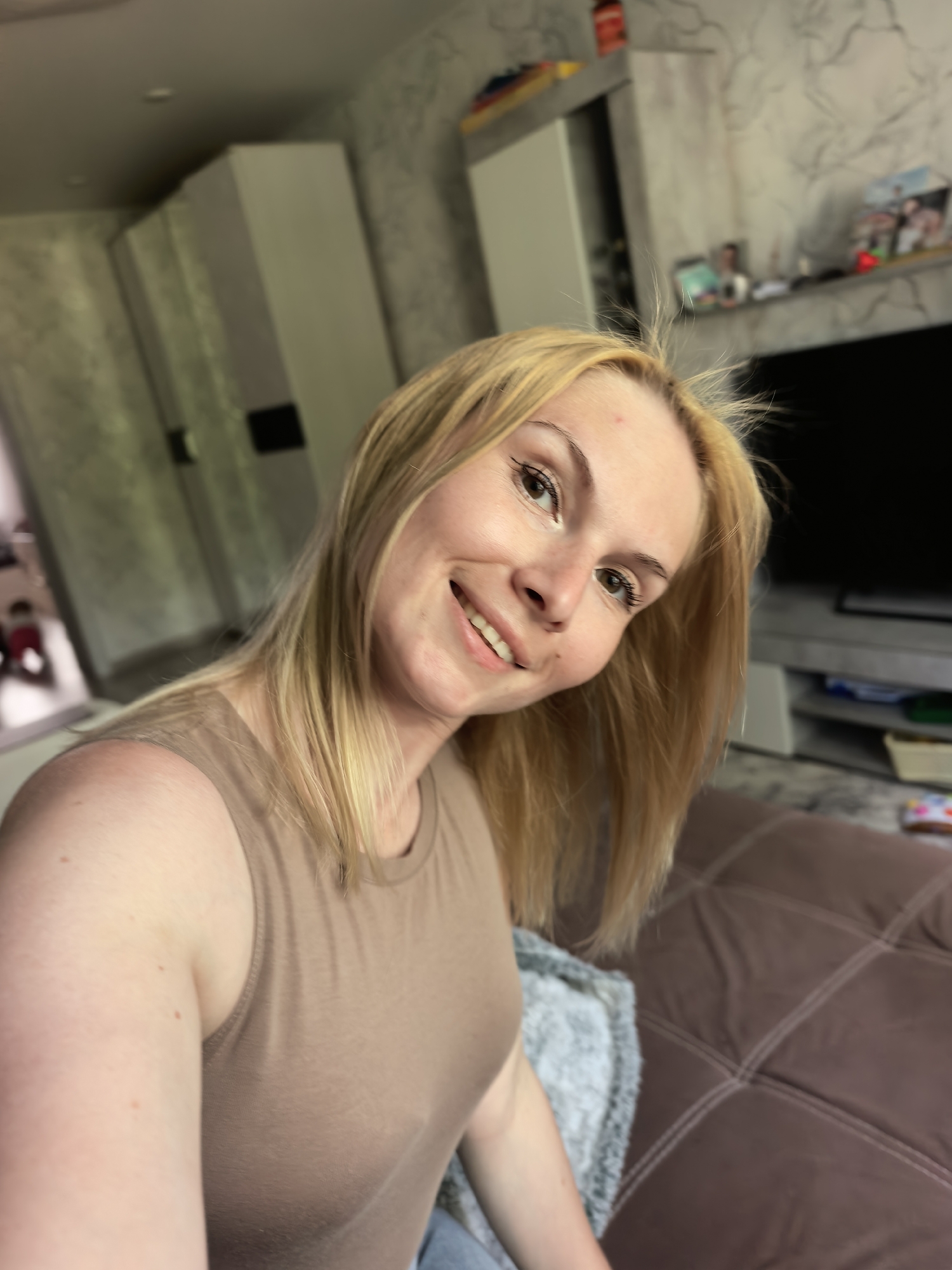 A woman with blonde hair smiles at the camera. She is wearing a tan tank top and is sitting on a brown couch with a grey blanket. In the background, there is a television, a shelf with pictures, and cabinets. The location is Russia, Sterlitamak.