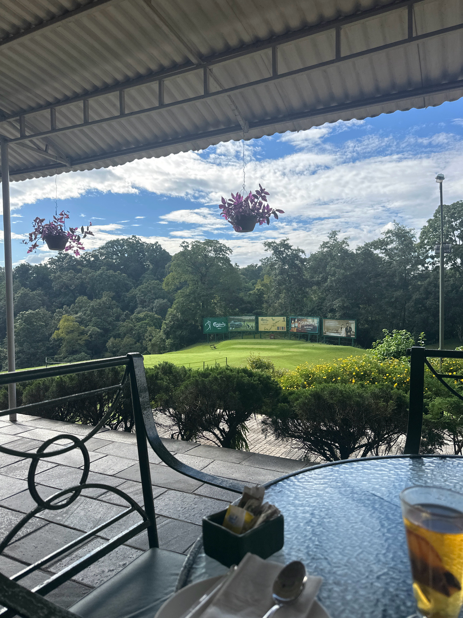The image captures a view from a covered patio overlooking a golf course in Kageshwori Manohara, Nepal. The patio features a metal railing and a table with a glass of amber liquid, a napkin, a spoon, and a small container of tea bags. In the background, a lush green golf course is visible, with trees lining the perimeter and a few advertising boards in the distance. The sky is a mix of blue and white clouds. Hanging flower baskets add a touch of color to the scene.