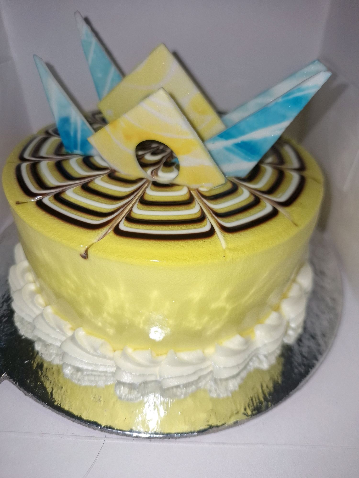 A yellow cake with white and blue chocolate decorations sits on a silver cake board in a white box. The cake is frosted with smooth yellow icing and decorated with a swirl pattern of brown, white, and yellow icing on top. It is adorned with triangular chocolate pieces, some of which are blue and white marbled, and others are yellow and white marbled. White frosting is piped around the base of the cake. The background is a plain white surface, suggesting an indoor setting. No people or specific city details are visible in the image.