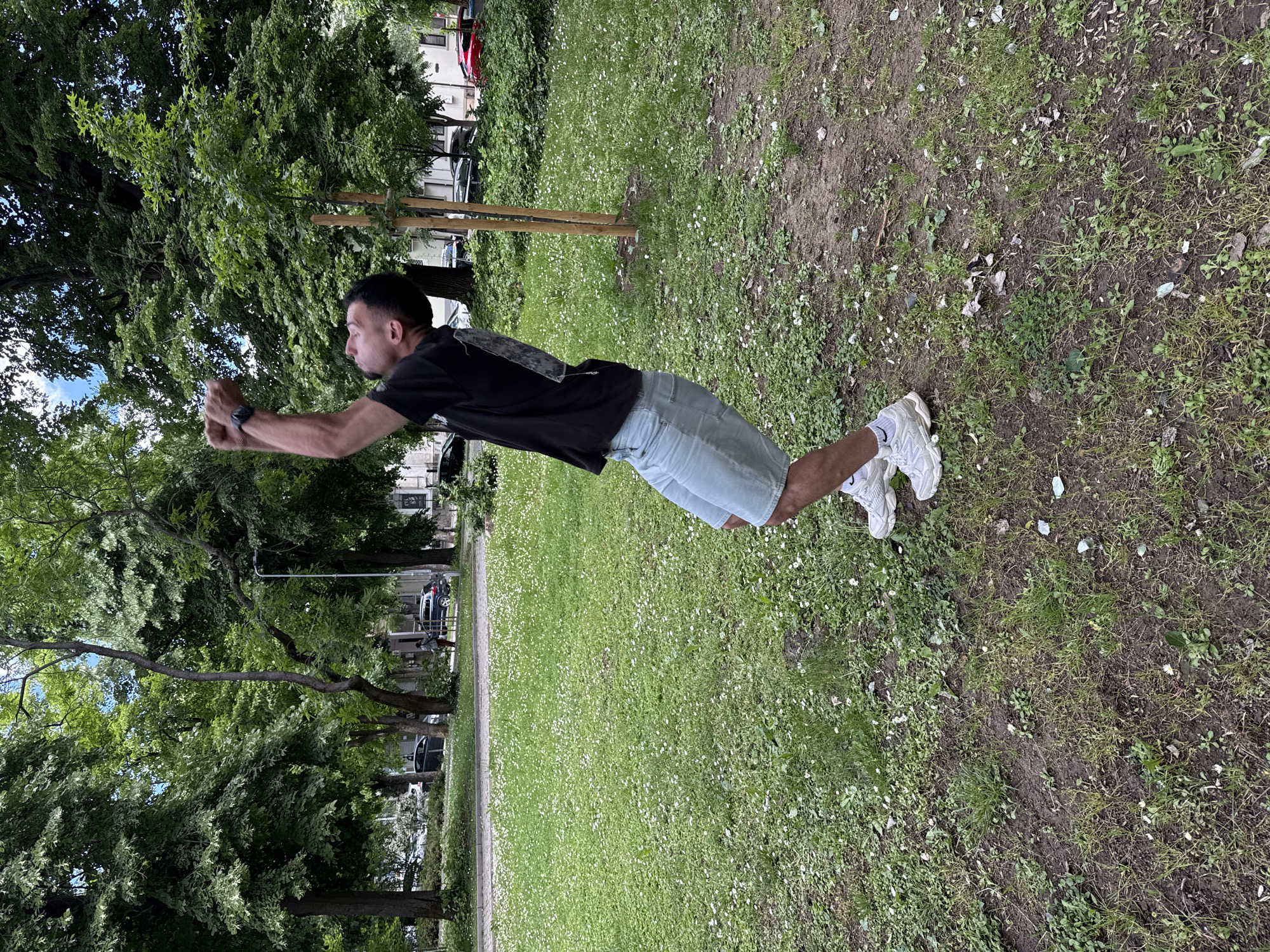 A man appears to be in mid-air, parallel to a grassy surface scattered with small white flowers. He wears a dark t-shirt, light-colored shorts, and white sneakers. His arms are extended forward, as if reaching for something. The background shows a park-like setting with trees, a building, and vehicles. The image appears to be taken near Veliko Tarnovo, Bulgaria.