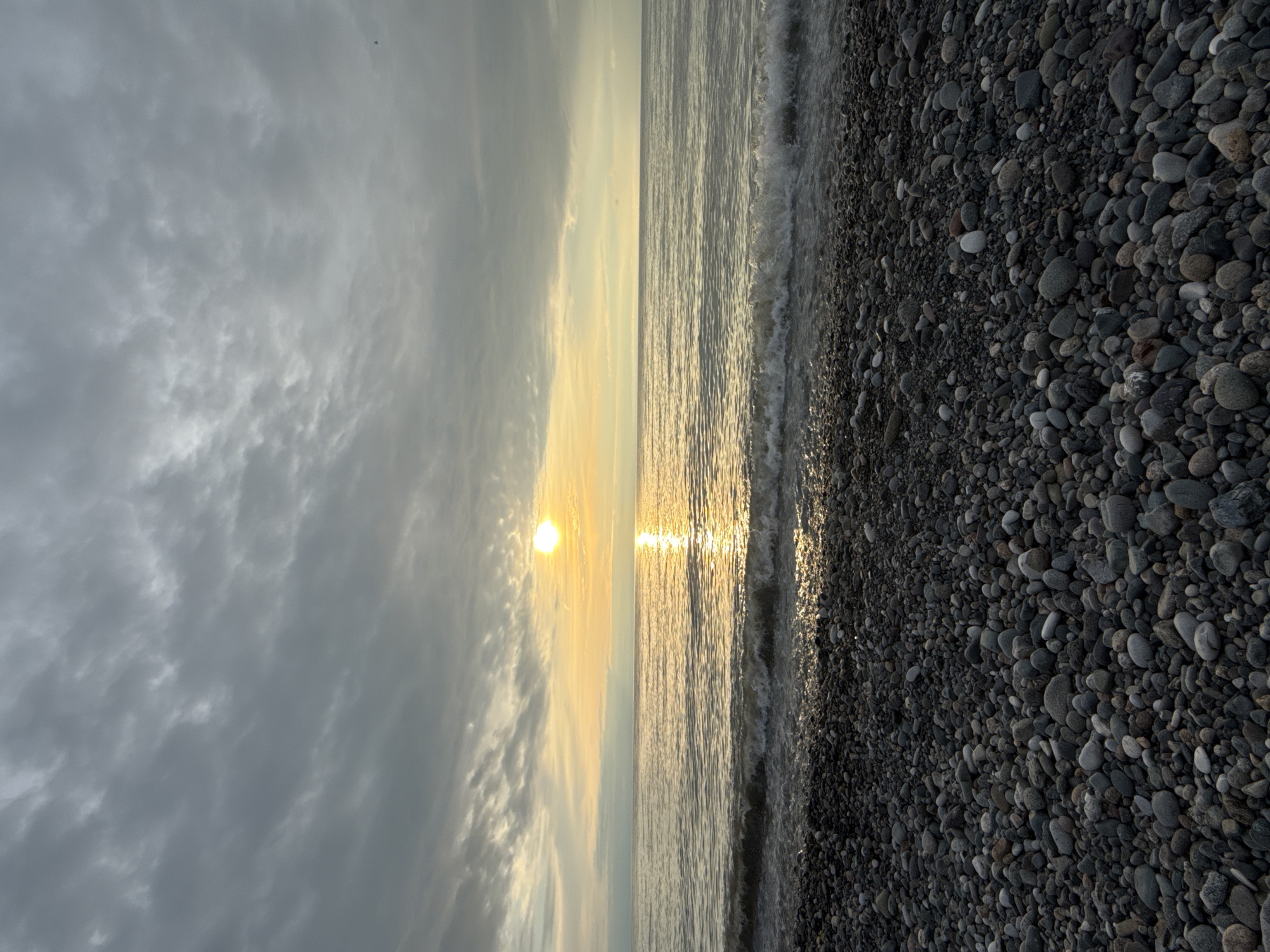 The image shows a vertical view of a beach scene, likely at sunset or sunrise, in Batumi, Georgia. On the left, heavy, grey clouds fill the sky. In the middle, the sun is visible, reflecting strongly on the water surface. On the right, there's a beach with small, dark stones. The ocean waves are gently lapping at the shore. No people are visible in the shot.