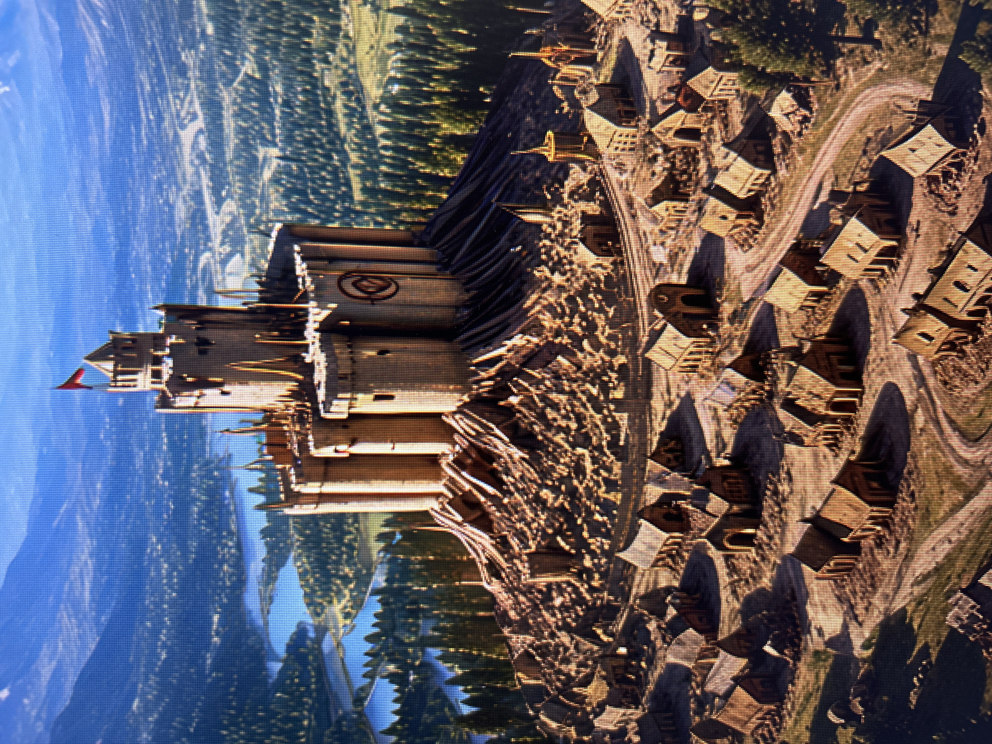 An aerial view depicts a medieval fortress situated atop a rugged hill, overlooking a vast landscape. The fortress, constructed from weathered stone, features multiple circular towers and battlements, with a red flag prominently displayed on its highest point. The hill itself is a chaotic jumble of debris and rough terrain, suggesting a history of conflict or construction. Surrounding the base of the hill is a village of simple wooden structures with thatched roofs, arranged along winding dirt paths. Beyond the village, the landscape opens to reveal a lush forest and a series of serene blue lakes, all framed by distant, mist-covered mountains. The overall impression is one of a powerful, isolated stronghold overseeing its domain.
