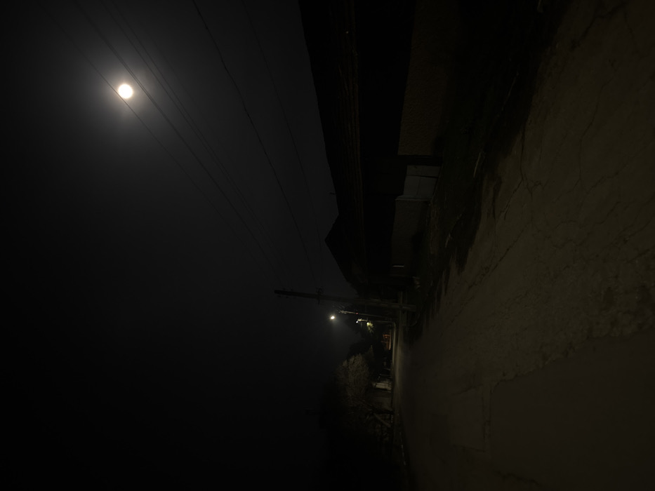 The image depicts a nighttime scene in Maslarevo, Bulgaria. A dirt or gravel road, marked with cracks and visible wet patches, extends from the foreground into the distance, gently curving left. On the left side of the road, sparse, bare trees or bushes are silhouetted against the dark. Further down this path, a second, fainter streetlight emits an orange glow. On the right side of the road, a utility pole with a bright white-yellow streetlight stands prominently, illuminating the immediate road surface and the facade of an adjacent low-slung building. This building features a dark, sloped roof and a lighter-colored wall or fence segment with a light-toned gate or door. Overhead, power lines traverse the dark sky, beneath a prominent, bright moon positioned in the upper right quadrant, appearing slightly diffused by atmospheric conditions. No individuals, animals, or vehicles are present.