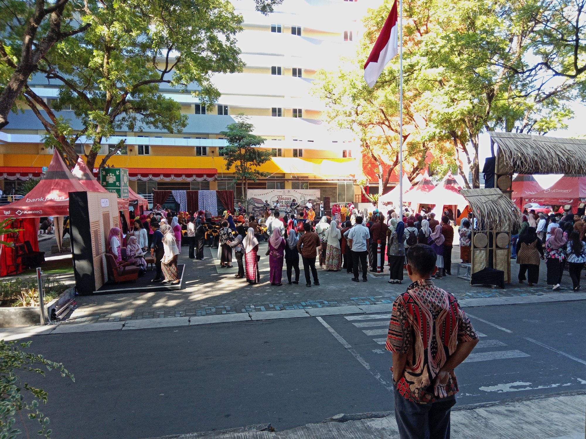 The image captures an outdoor event, likely a bazaar or festival, in front of a modern multi-story building. The architecture of the building suggests it could be a university or office complex, with multiple floors and evenly spaced windows. A prominent Indonesian flag, red and white, waves from a flagpole on the right side of the frame, indicating a national celebration or event.
In the foreground, a man stands with his back to the camera, wearing a batik shirt and dark pants. He is looking towards the event. The ground is paved with asphalt and marked with a pedestrian crossing.
The middle ground reveals a bustling scene with many people dressed in traditional Indonesian attire, including batik and headscarves. Several stalls or booths are set up, some covered with thatched roofs and others with red canopies. One canopy has the "bankjatim" logo visible, suggesting a sponsor. There are decorative elements like bunting and some furniture such as chairs and a sofa displayed. Trees with lush green foliage provide shade, and dappled sunlight creates patterns on the ground.
In the background, the large building dominates the skyline. The event appears to be lively, with people mingling and interacting. The overall atmosphere is festive and communal. While the specific location in Malang, Indonesia, is not explicitly stated, the presence of the Indonesian flag and the style of dress strongly suggest it is within the country. The architectural style of the building could be characteristic of modern Indonesian urban development.