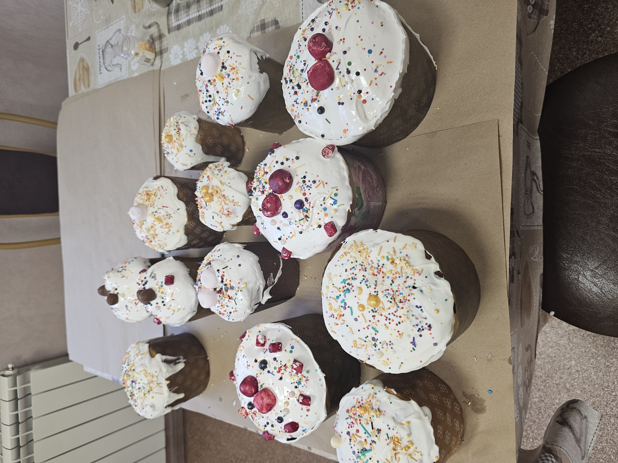 The image shows a tabletop arrangement of eleven Easter cakes ("kulich") decorated with white icing and colorful sprinkles. The cakes vary in size, with some topped with candied cherries or marshmallows. They rest on a sheet of brown paper, likely for cooling or display. In the background, there is a partially visible tablecloth with a patterned design, a radiator, and a glimpse of a dark leather chair. The setting is likely indoors in a private residence. No people are visible. There is no indication of Moscow, Russia.