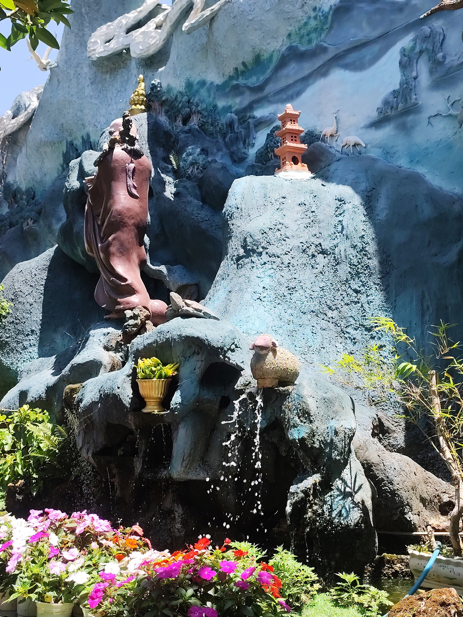 The image captures a Buddhist shrine in Xuân Thành, Vietnam. A reddish-brown statue of a monk stands prominently on a blue-gray rock formation. Additional small statues, including a golden Buddha, a three-tiered pagoda, and a sheep, are placed on the rock. The rock structure has built-in water features, with water cascading down into a small pond surrounded by potted colorful flowers. Green plants and small trees flank the rock structure. The background includes a painted mural of mountains and deer.
