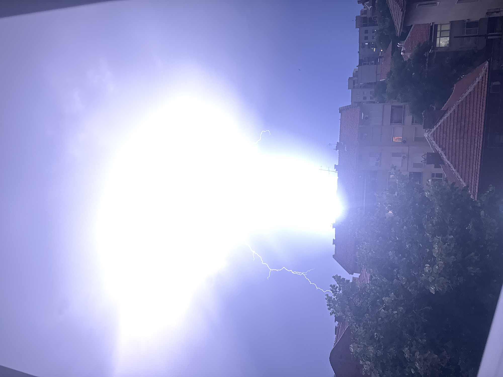 The image captures a dramatic lightning strike illuminating the sky over a residential area in Plovdiv, Bulgaria. The bright flash emanates from the upper center of the frame, casting an intense light that washes out details in the sky and creates lens flare. A jagged lightning bolt is visible extending downwards from the bright core. Below the illuminated sky, the silhouettes of buildings with red-tiled roofs are partially visible, some with lit windows suggesting indoor activity. Trees with dark foliage are present in the foreground, obscuring some of the lower structures. The overall scene depicts a powerful natural phenomenon occurring within an urban environment.