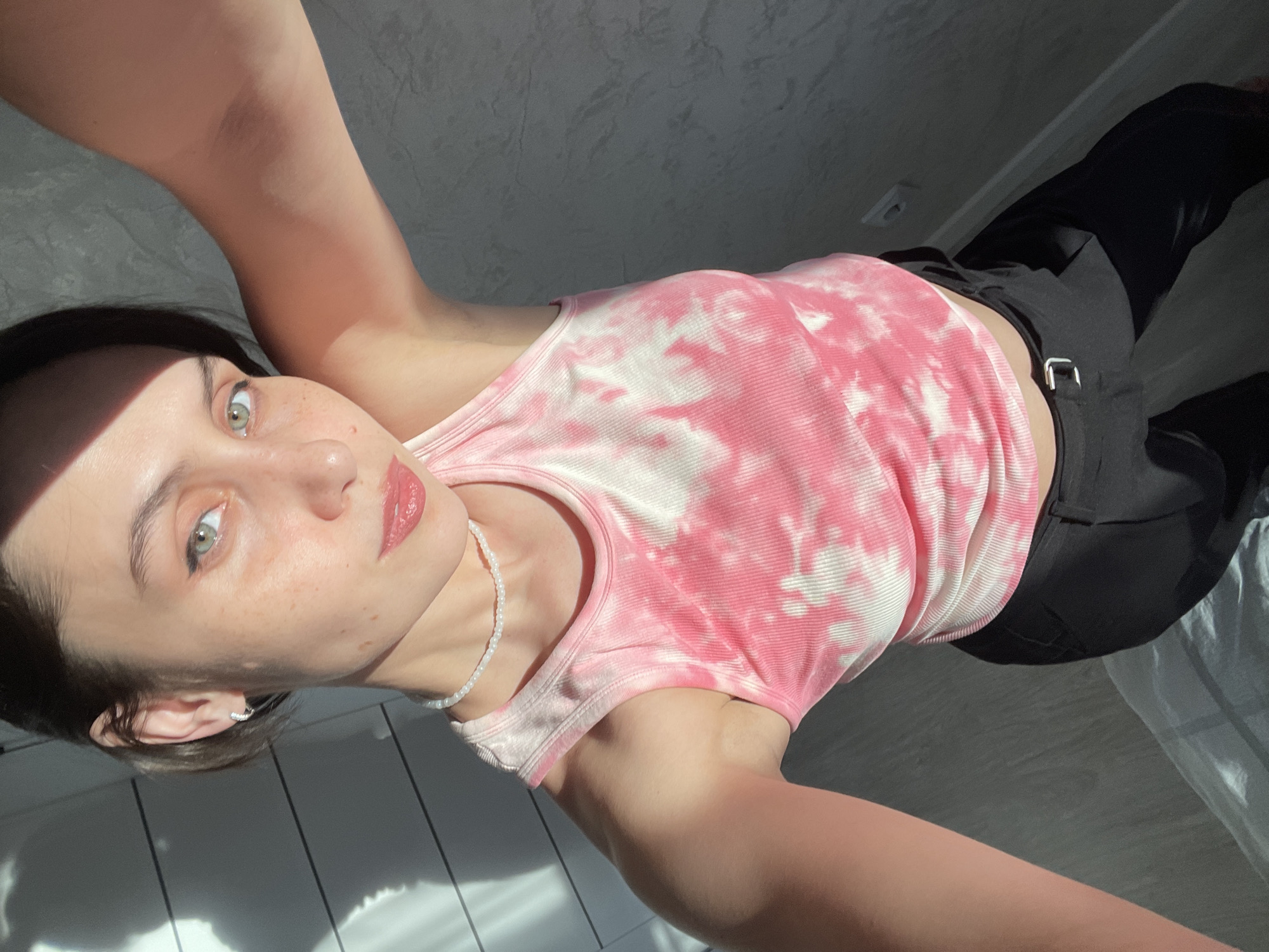A young woman is visible in a selfie taken at an upward angle. She has fair skin, freckles, light green eyes, and dark hair pulled back. She is wearing a pink and white tie-dye tank top and black pants. A white beaded necklace is around her neck. Her location in the city of Izhevsk, Russia cannot be determined from this image. She is standing in a room with light gray tiled flooring and a light gray wall. Sunlight is streaming into the room, casting shadows across the floor and her face.