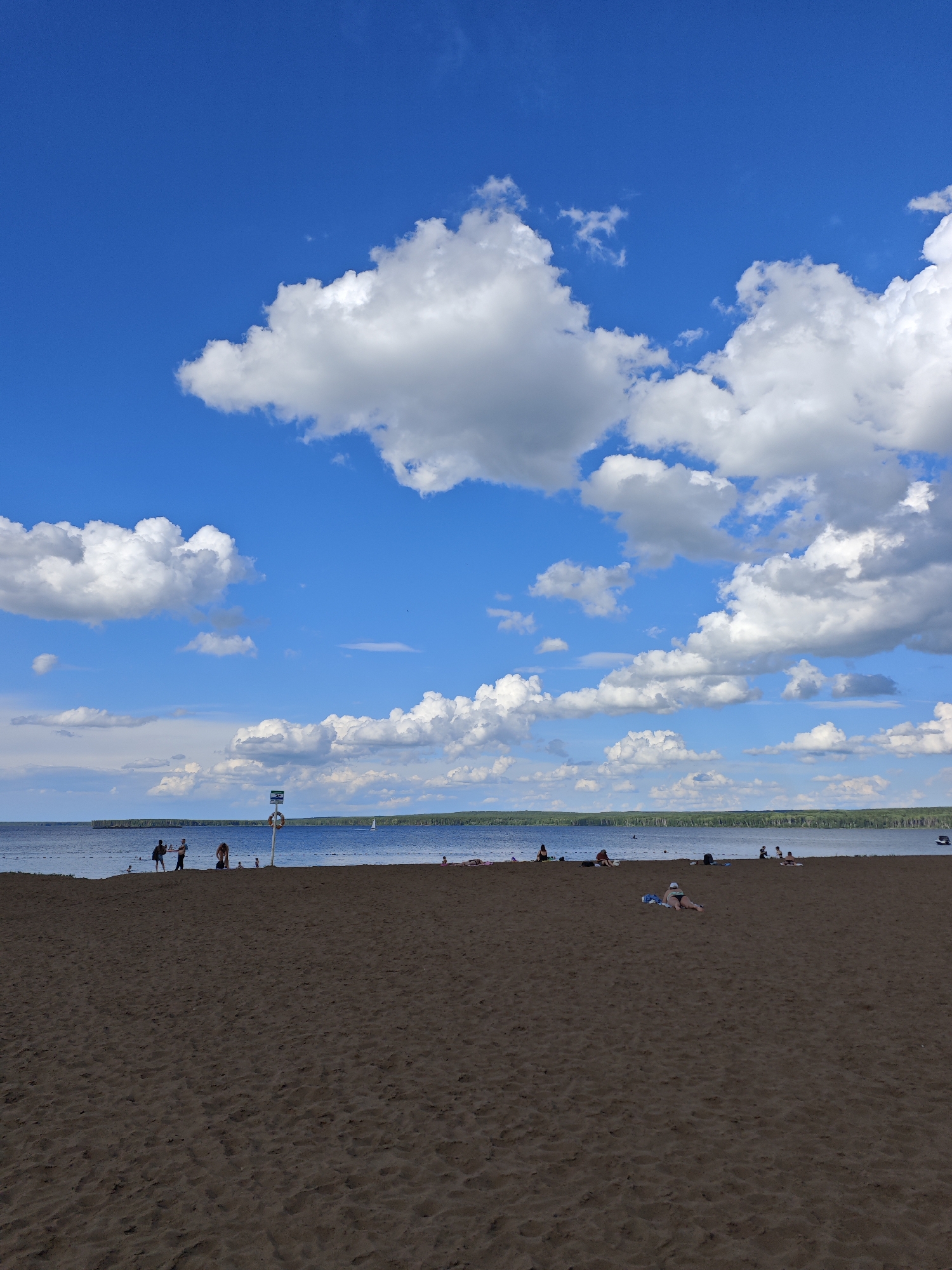 This is a vertical, eye-level, full shot of a beach scene on a sunny day. The sky is a brilliant blue, populated by numerous fluffy white clouds. In the foreground, a wide expanse of dark brown sand stretches out, marked with the textured patterns of footprints and the gentle ripples left by the wind. The middle ground features the blue water of a lake, with a distant treeline forming the horizon. Several people are scattered across the sand and near the water's edge. On the beach, individuals are walking, sitting, and lying down. Near the water, some people are standing and interacting. A life preserver ring on a pole is visible on the sand near the waterline, indicating this is a designated swimming area. The beach is part of the Kama River embankment in Chaikovsky, Russia, a popular recreational spot for locals and visitors.