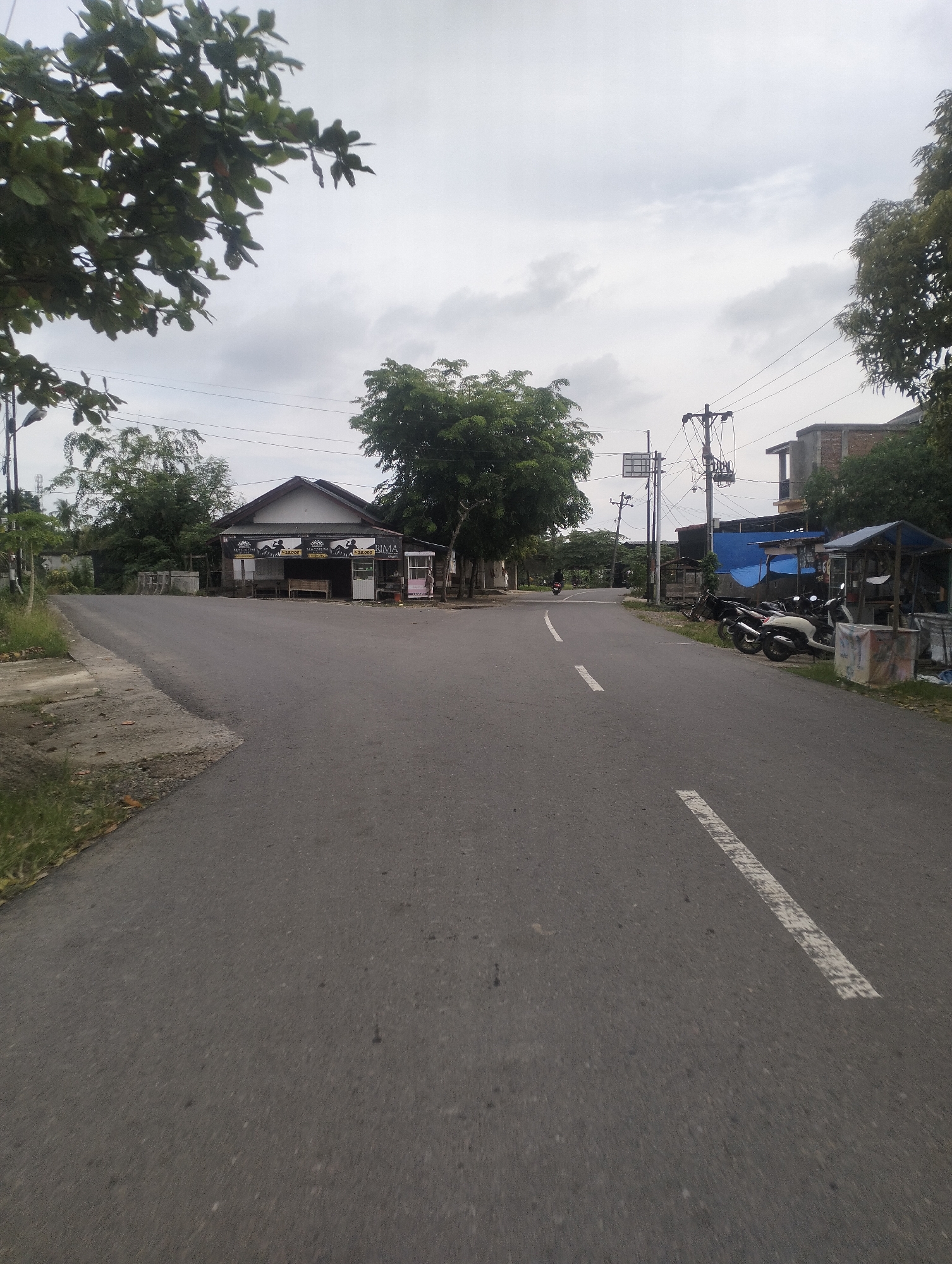 The image shows a street scene in Muliengmanyang, Indonesia, on an overcast day. A paved road curves from the foreground towards a small cluster of buildings on the left and right sides of the road. The building on the left appears to be a small shop with a peaked roof and a sign that reads "RIMA". Several motorcycles are parked in front of structures on the right side of the road, one with a blue tarp roof. A single motorcycle is visible in the distance on the road, heading away from the viewer. Trees and utility poles are scattered throughout the scene. The sky is heavily clouded.