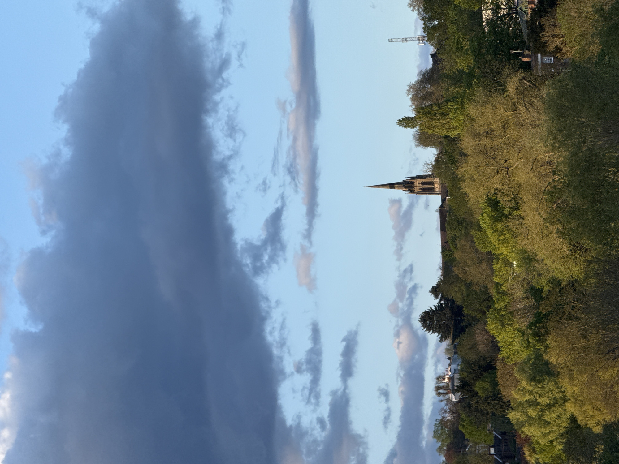 The image shows a landscape in London, United Kingdom, with a church steeple and tree line visible against a sky with various cloud formations. A construction crane is visible above the treeline. The sky is a mix of blue and dark grey clouds. Buildings and structures are partially obscured by trees.