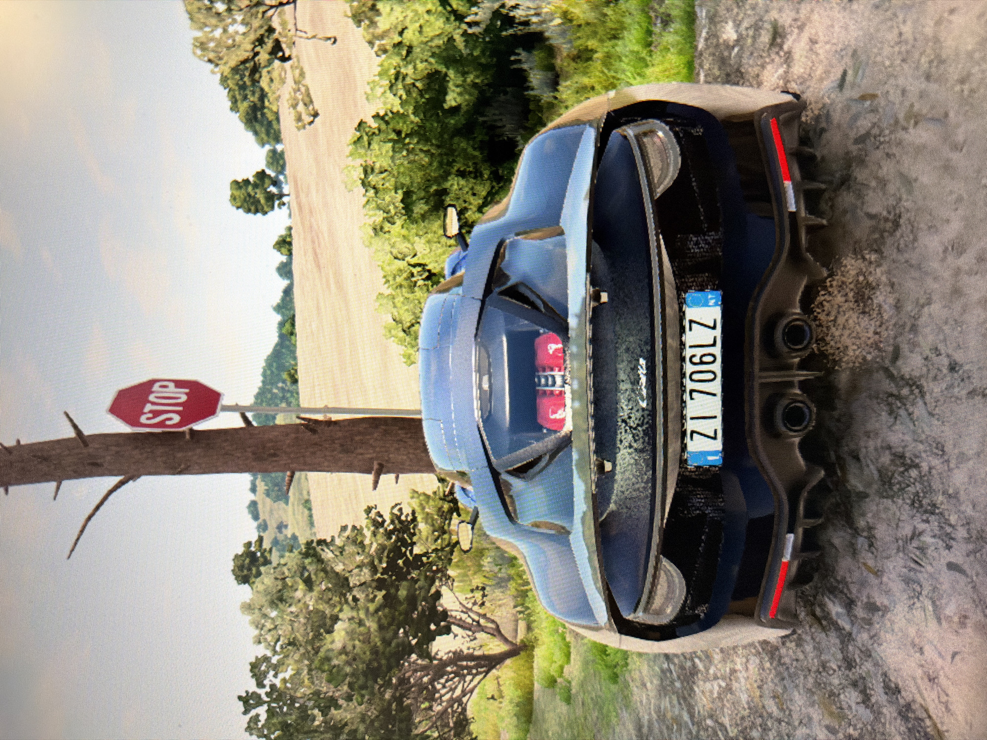 An image, rotated 90 degrees counter-clockwise, displays the rear view of a blue sports car on what appears to be a dirt or gravel road. The car has a sleek design with red engine components visible through a rear window or vent, dual exhaust pipes, and red taillights. A European-style license plate is clearly visible on the back, displaying the text "ZI 706LZ".
To the left of the car, a red octagonal "STOP" sign is mounted on a rough wooden post or tree trunk. The background features a bright sky, green trees, and an open field or rolling hills under what seems to be daylight conditions. The overall scene suggests a rural or countryside setting, possibly in a warm climate. The image exhibits a pixelated quality, indicative of a screenshot from a video game. No people are visible.
