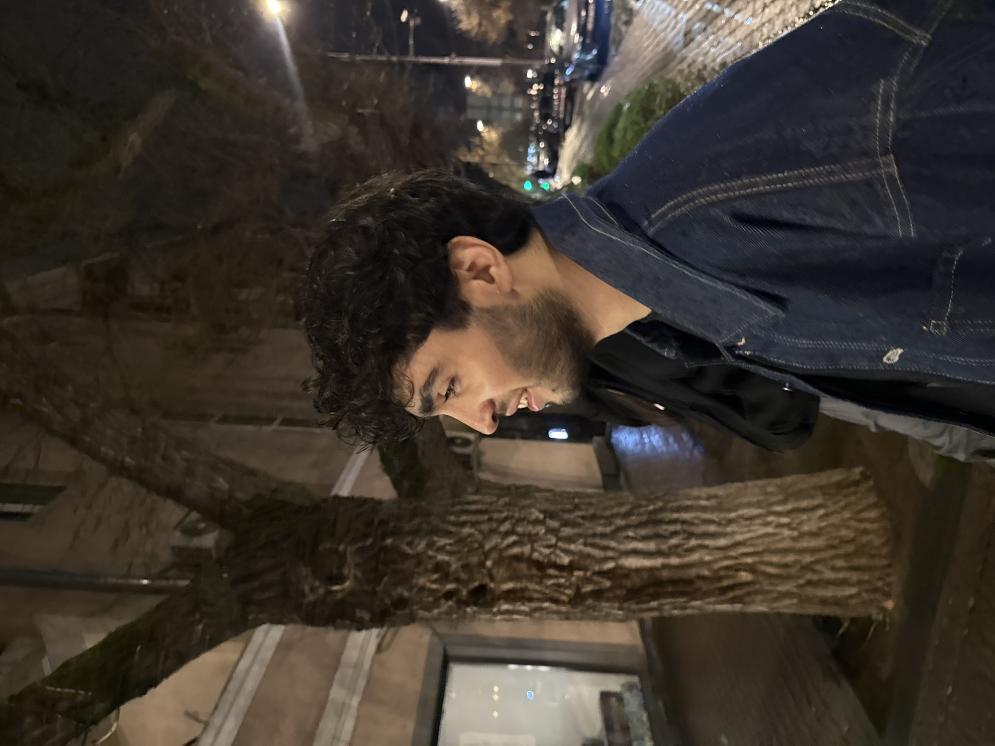 A dimly lit, tilted, outdoor nighttime shot depicts a man with dark hair and a beard. He is wearing a denim jacket and a black shirt. His face is slightly illuminated, and he appears to be smiling or talking. In the background, there's a tree trunk and branches. Beyond the tree, one can see city lights, likely from street lamps and vehicles. Based on the environment, this image appears to have been taken in Yerevan, Armenia. A window or storefront with a light source is visible behind the tree trunk. The ground appears to be paved.