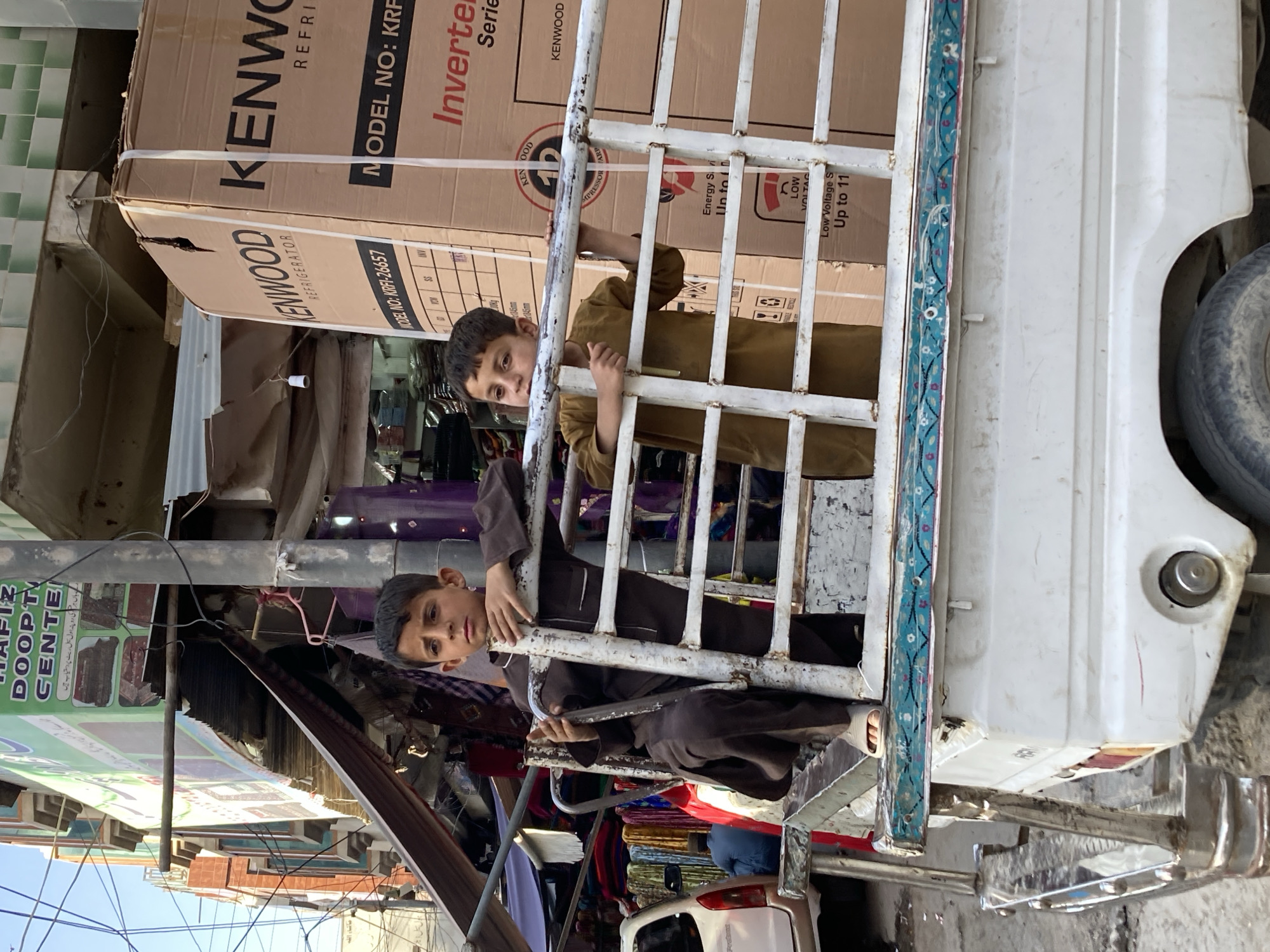 The image captures a vibrant street scene, likely in a market area of Quetta City Tehsil, Pakistan, under bright daylight. Two young boys are prominently featured, peering out from what appears to be the back or side of a white utility vehicle, possibly a small truck or rickshaw. Both boys, dressed in traditional attire, are holding onto the white metal grate that forms part of the vehicle's enclosure, looking directly at the camera with curious expressions. The grate features a decorative blue and pink floral pattern along its top edge.
Behind the children, large cardboard boxes are stacked, clearly labeled "KENWOOD REFRIGERATOR." Specific text visible on these boxes includes "MODEL NO: KRFJ-26657," "Inverter Series," "Energy Saving," and "Low Voltage Start Up to 11."
The background reveals a bustling market environment. To the left, a building with green and white tiled walls is visible. Below the vehicle, various market goods, possibly colorful textiles, and parts of a white car can be glimpsed. Further into the background, utility poles and numerous power lines crisscross the sky, and a large green banner is displayed. Text on the banner includes "ماشاء اللہ," "HAFZ DOOP'TY CENTER," and Urdu script that translates to "All kinds of carpets are available here at first hand and at wholesale rates." The overall scene depicts a moment of daily life in a busy urban setting.