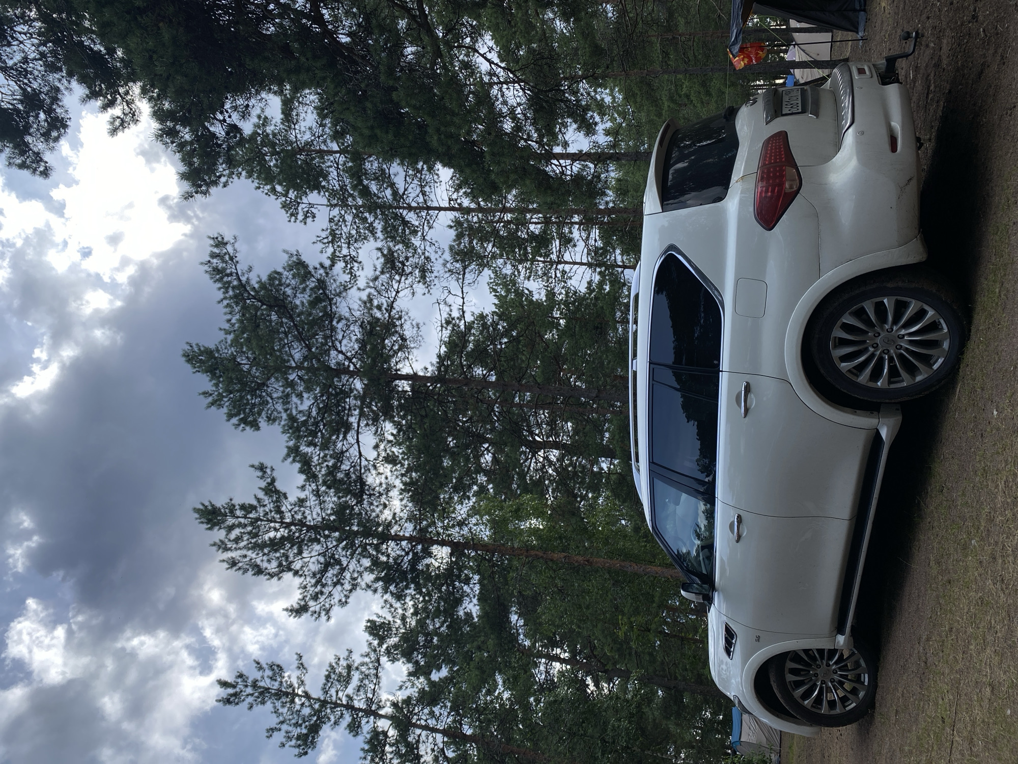 This media file is a photograph of a white SUV parked on a dirt path in a forest. The car is positioned diagonally in the frame, with its rear to the right. Pine trees with dark green needles surround the scene, and the ground is covered in dry grass and pine needles. The sky above is cloudy and overcast, suggesting a possibly rainy or stormy day.
In the background, partially visible to the right of the SUV, are two tents and what appears to be camping equipment, indicating that the setting is a campsite. A license plate is visible on the rear of the car, but the text on it is not legible. The overall mood of the image is serene and rustic, conveying a sense of being in nature. The lighting is diffused due to the cloudy sky, and there are no people visible in the frame. The photo appears to have been taken during daylight hours, likely in the afternoon or evening given the subdued light.