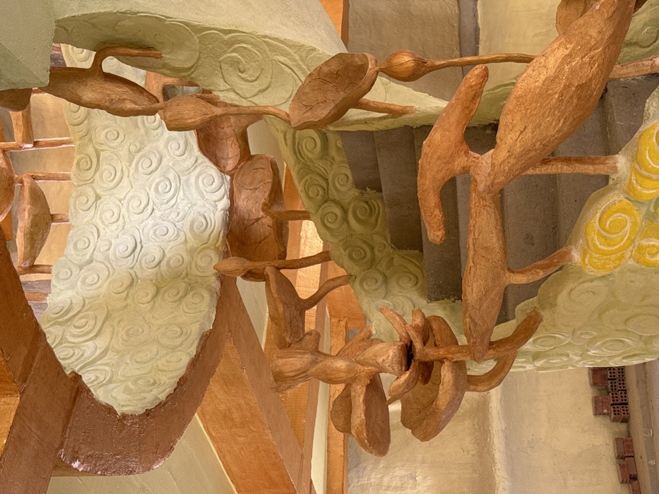 A decorative spiral staircase is depicted in an indoor or covered outdoor setting in Đăk Mar, Vietnam. The staircase features grey concrete steps. Its outer wall, serving as a balustrade, is light green with raised, swirling cloud patterns, bordered by a brown curved structure. The handrail is sculpted in an organic, branch-like form and painted a metallic gold-bronze. Numerous sculpted elements resembling lotus leaves and lotus buds, matching the handrail's color, are attached to the handrail and extend upwards along the spiral. Background elements include cream or pale yellow walls and brown structural beams. A stack of reddish-brown bricks is visible on a light floor in the lower-left corner. No individuals or specific actions are depicted.
